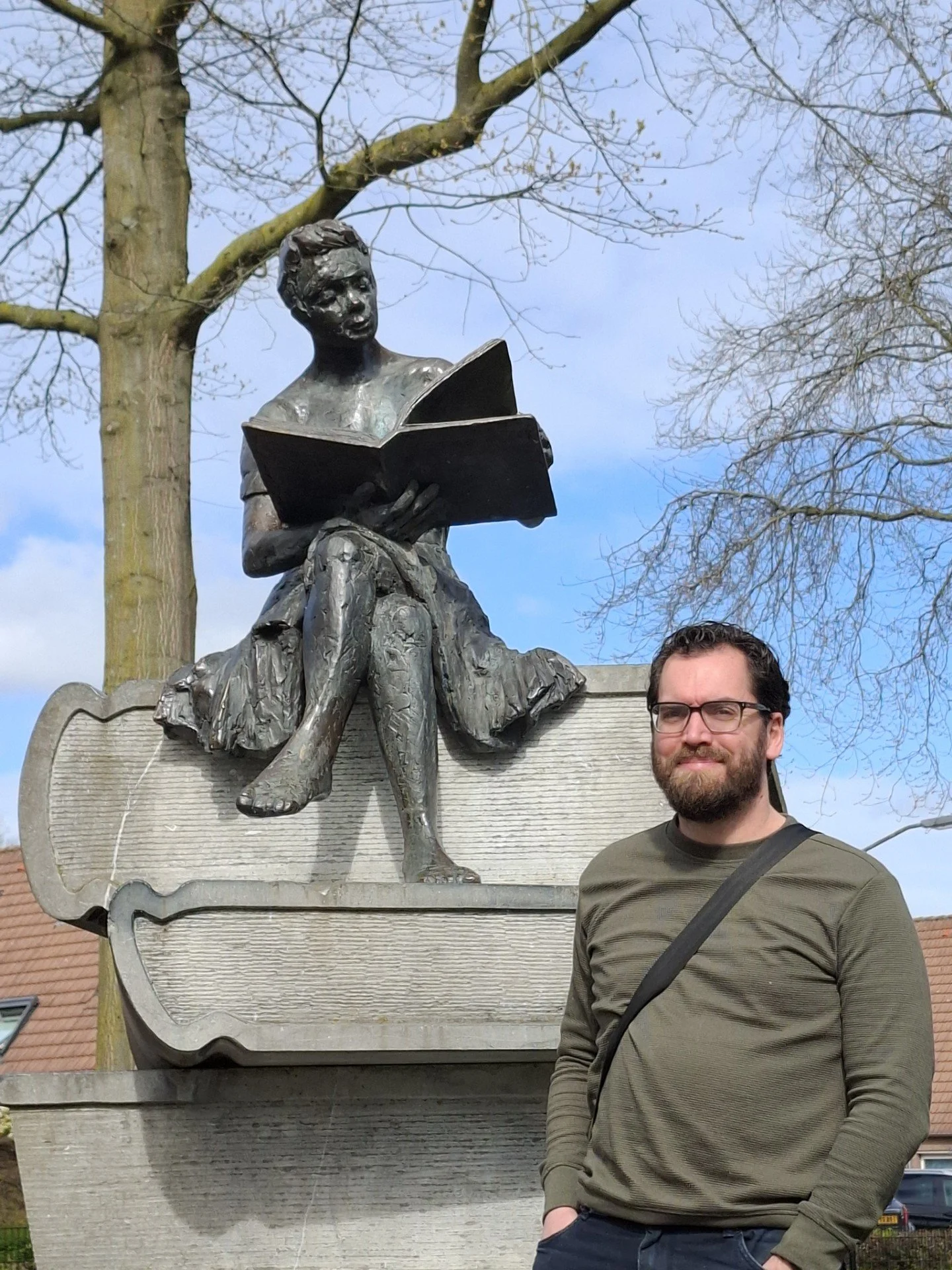 Een man met een bril en een baard staat naast een beeld van een kinderfiguur dat op een boek zit, in een park met bomen en een blauwe lucht.