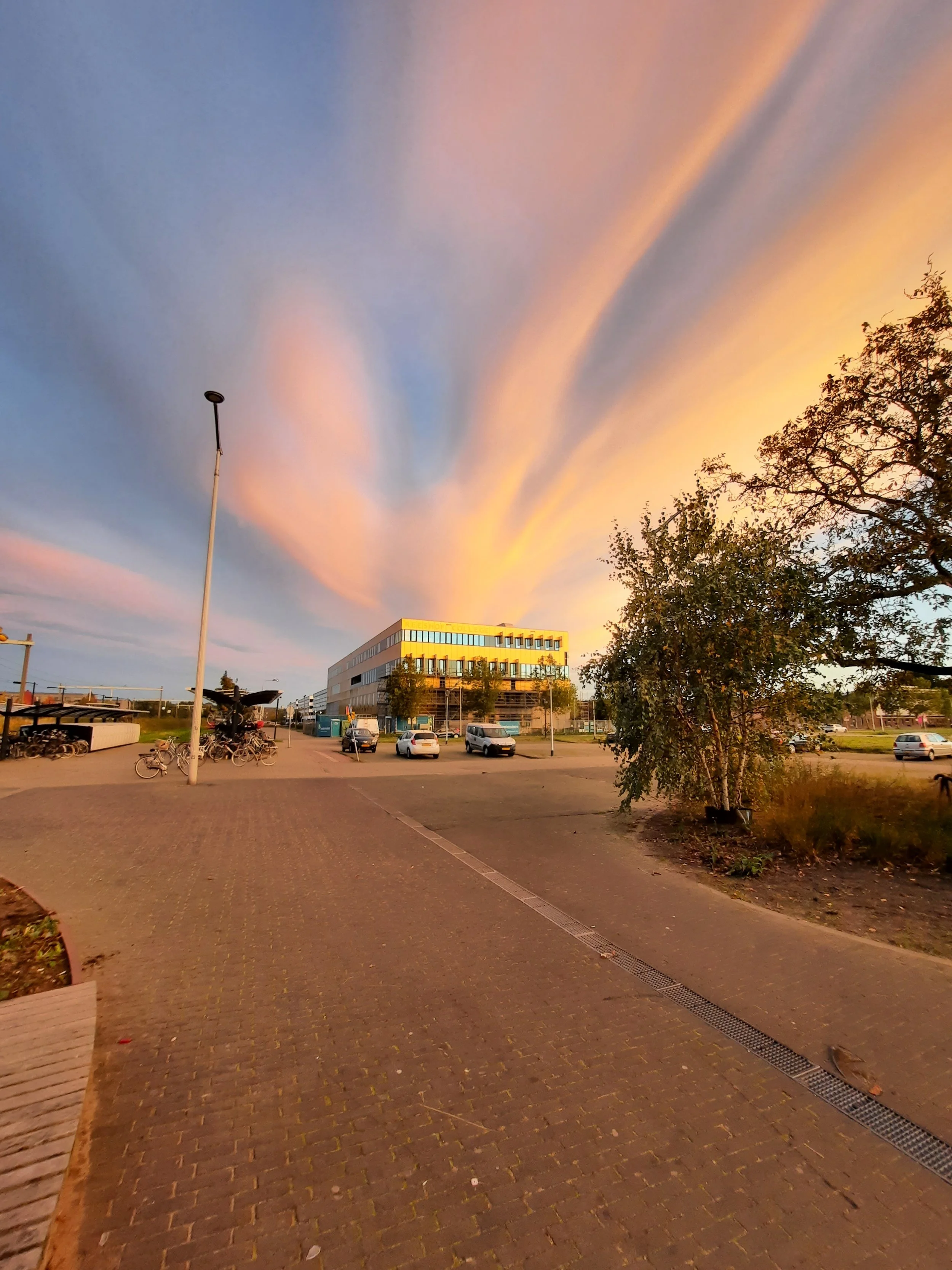 Een straat met een parkeerplaats, fietsen, een paar auto's en een modern gebouw onder een kleurrijke, bewolkte sky tijdens zonsondergang.