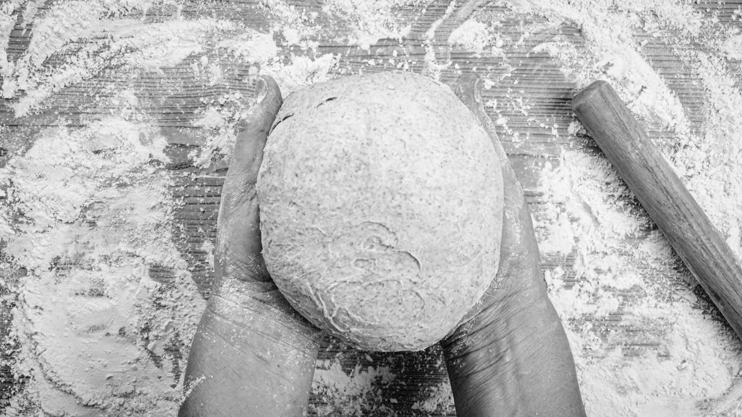 Dough ball held in a pair of hands on top of a flour covered wood counter