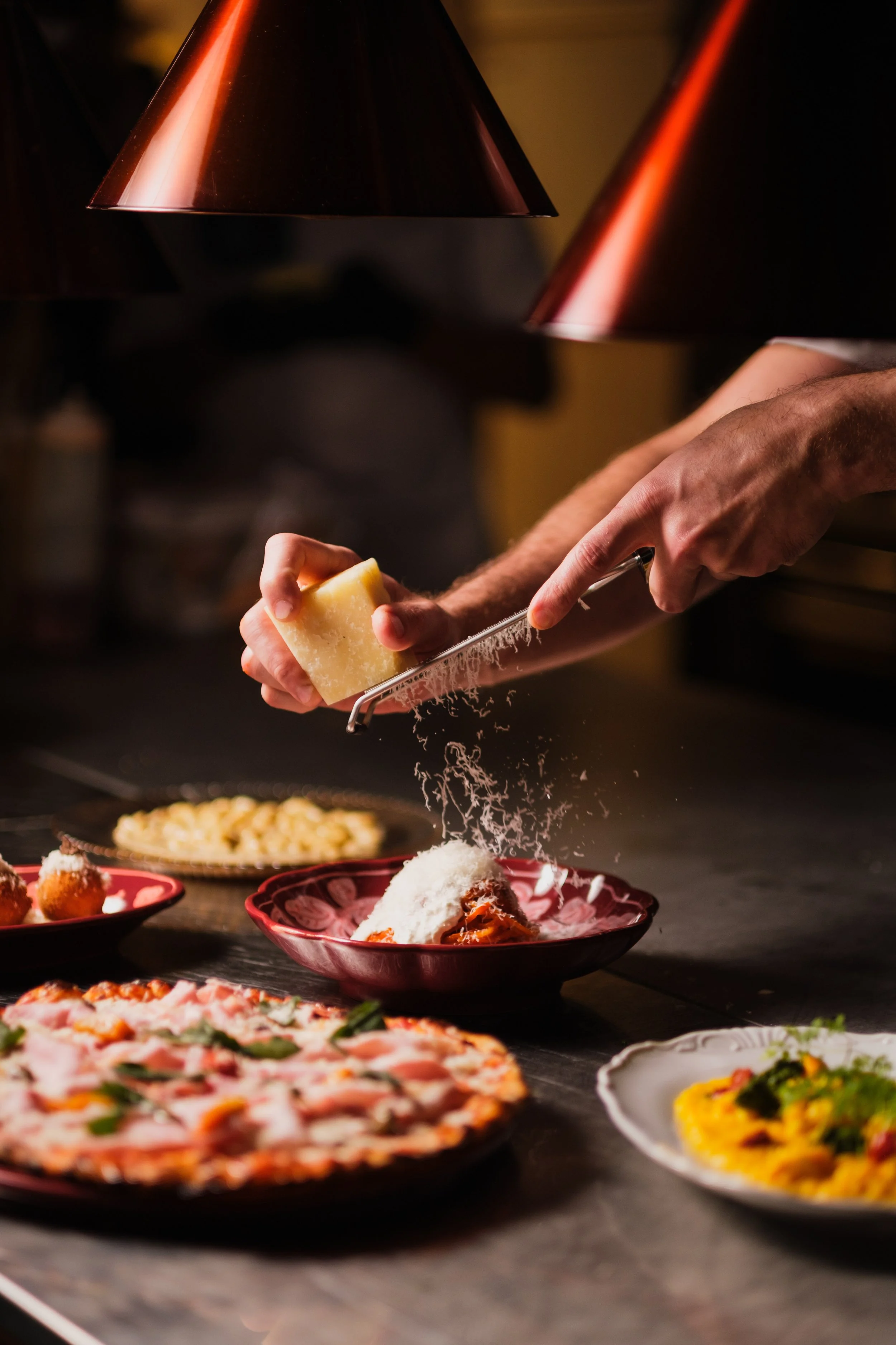 Närbild av en person som riven ost över en pizza, med andra rätter på bordet, som olika piroger och en pizza, i bakgrunden.