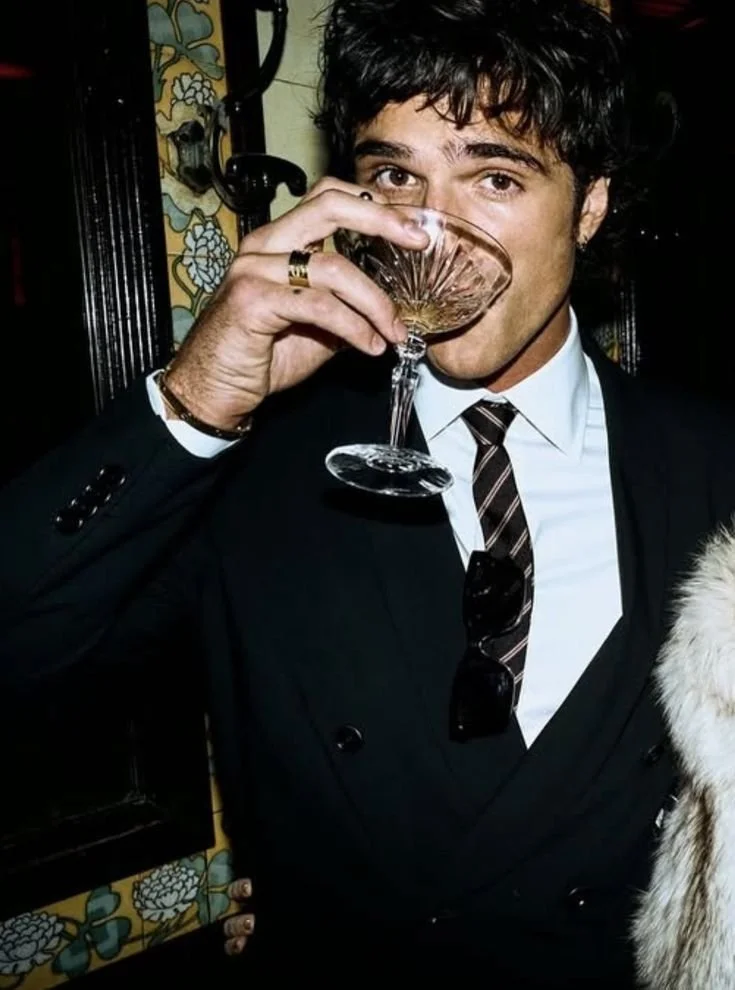 En ung man i vit skjorta, mörk kostym och slips som dricker från ett cocktailglas, troligtvis vin eller liknande, i ett inomhusmiljö.