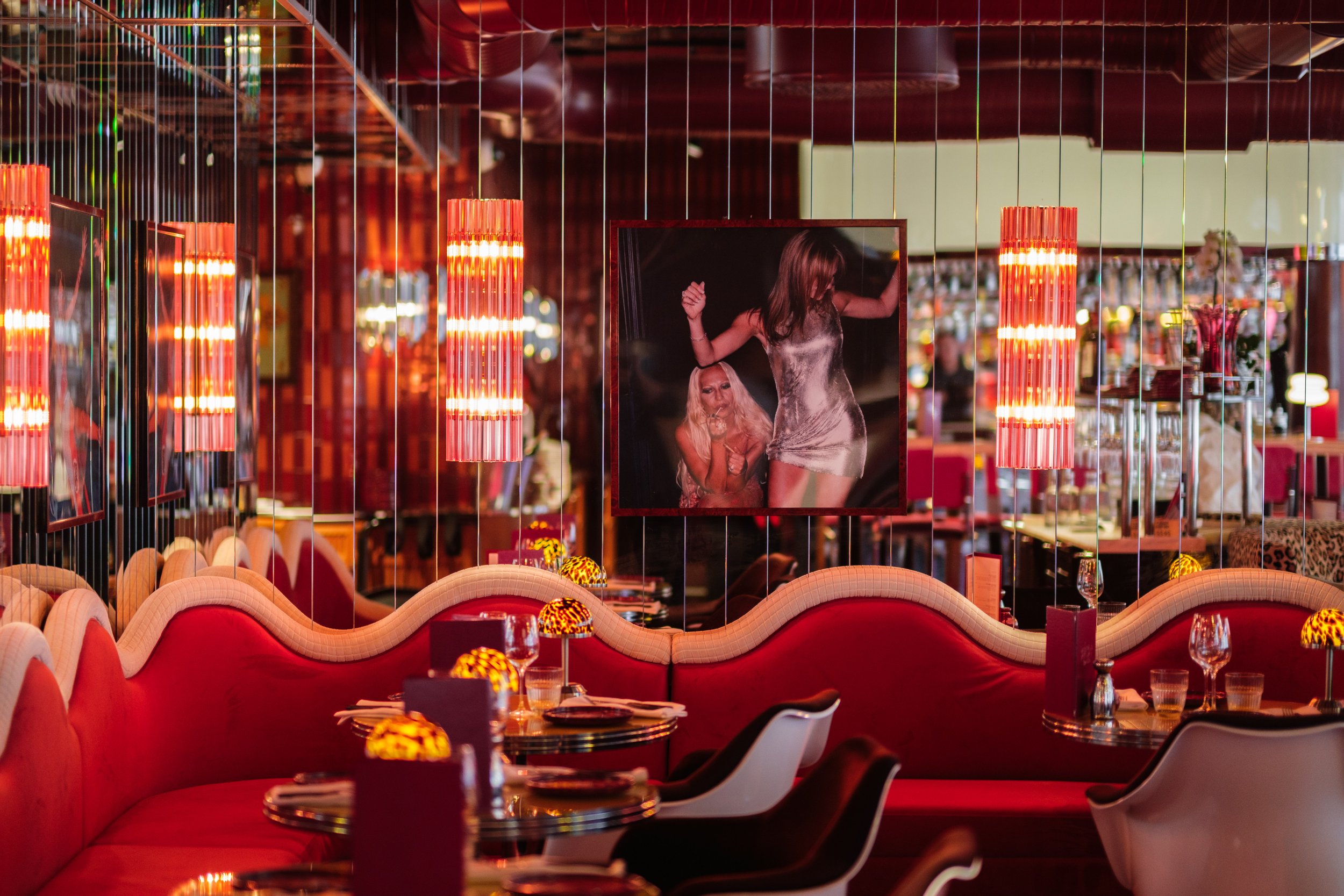 Inredning av en restaurang med röd och rosa färgtema, färgglada lampor, stora fotografier och runda bord med ljus, samt vita och svarta stolar.