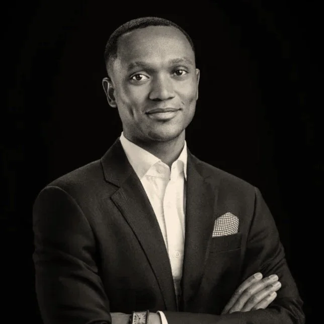 Black and white professional portrait of a man in a suit with arms crossed, smiling gently against a dark background.