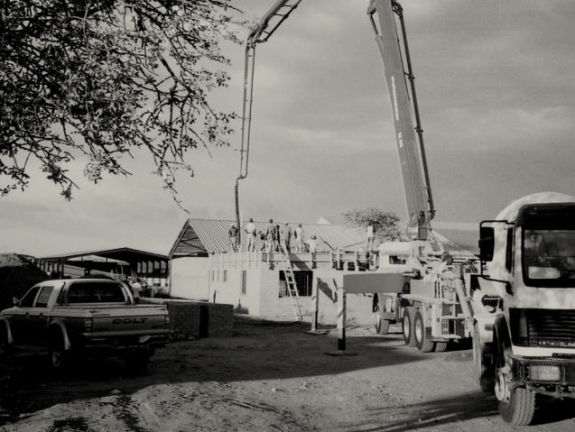 Construction site with a crane pouring concrete on a building under construction, with trucks parked nearby and a tree in the foreground.