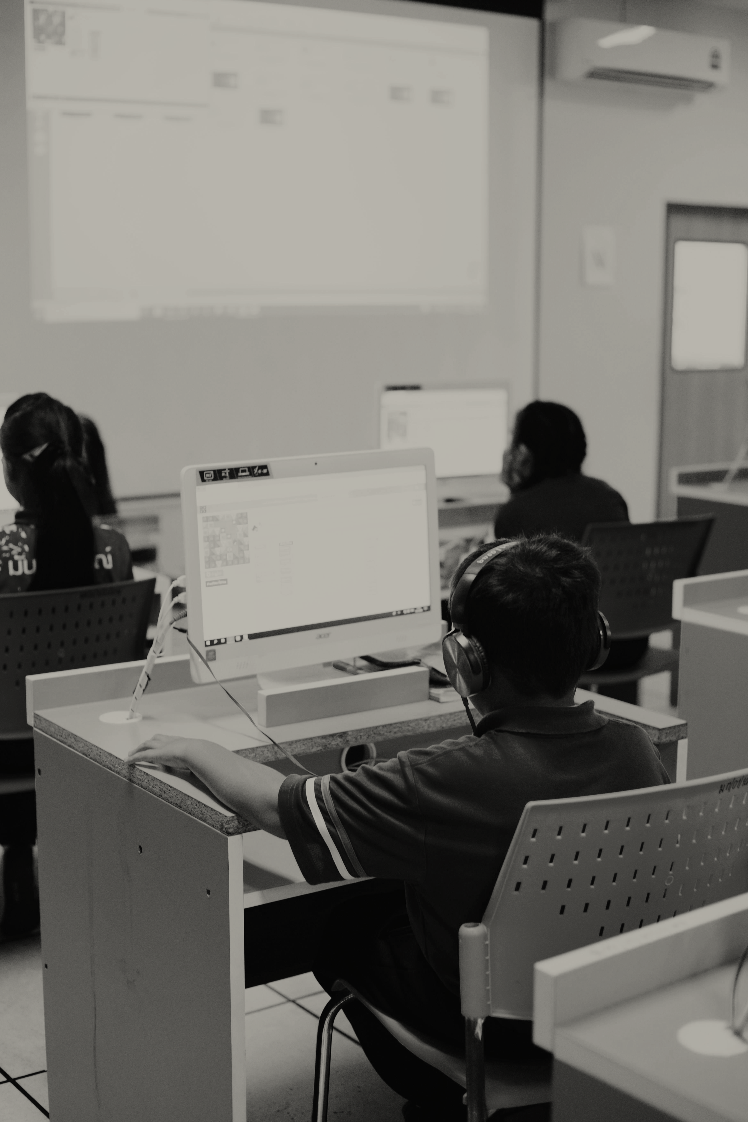 Students sitting at desks in a classroom with computers, listening to a lecture or presentation projected on a whiteboard.