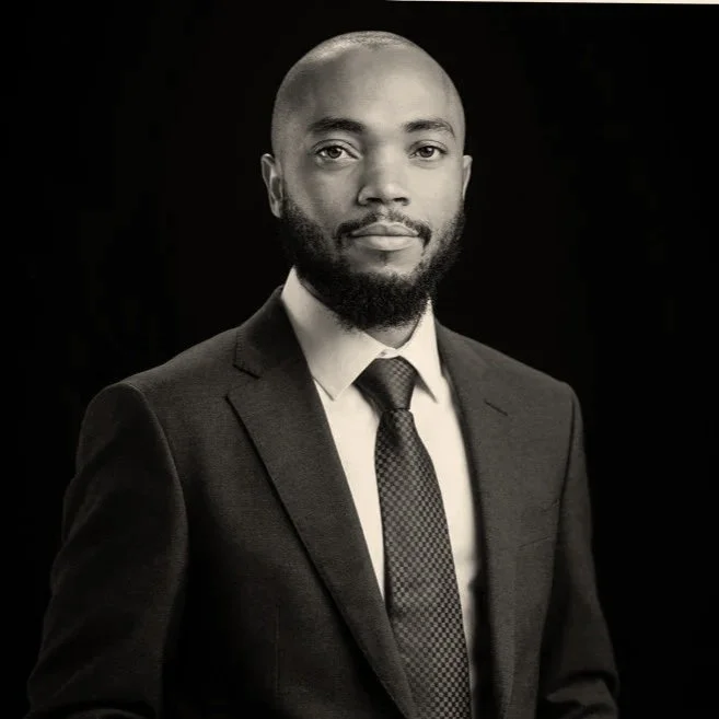 Black and white professional portrait of a man with a beard in a suit and tie against a dark background.