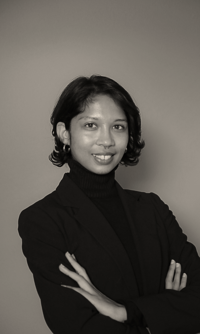Black and white portrait of a woman with short, wavy hair, wearing a dark turtleneck and blazer, smiling with arms crossed against a plain background.
