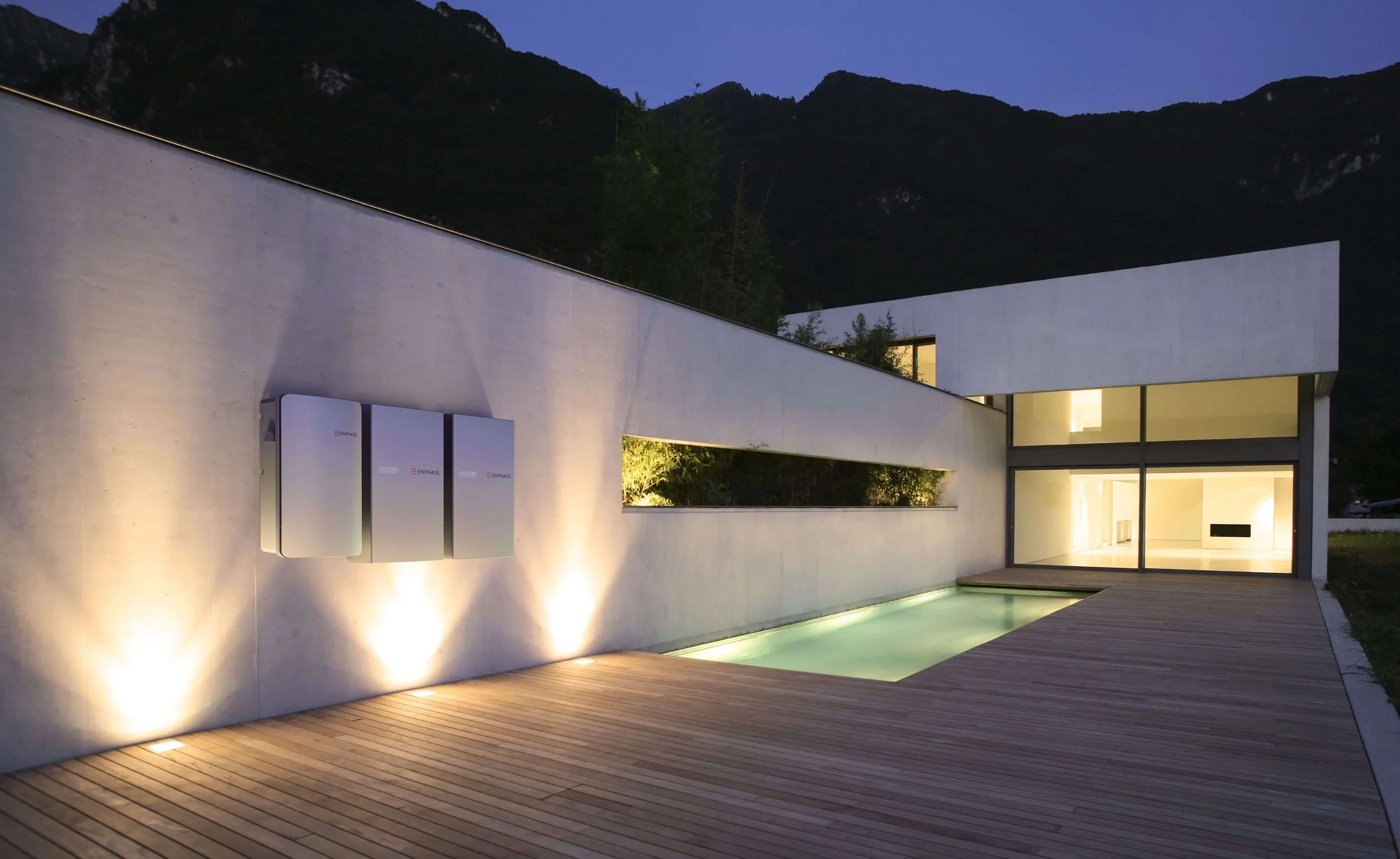 Modern house with Enphase controller and battery storage on outdoor deck, illuminated at night with mountain background.