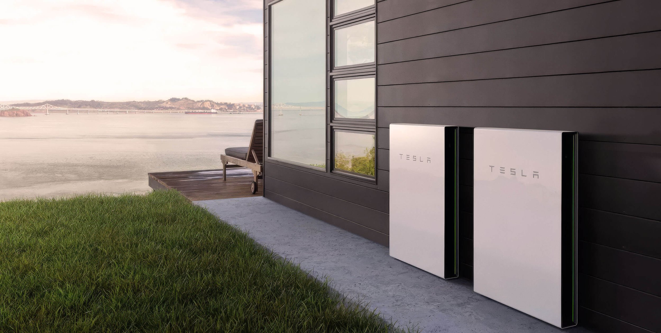 Modern house exterior with two white Tesla PowerWall batteries, and a waterfront view with a bridge in the distance.