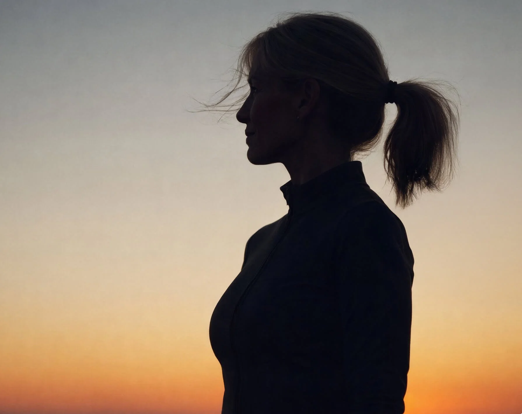 Silhouette of a woman with her hair tied back in a ponytail, facing left, during sunset or sunrise.