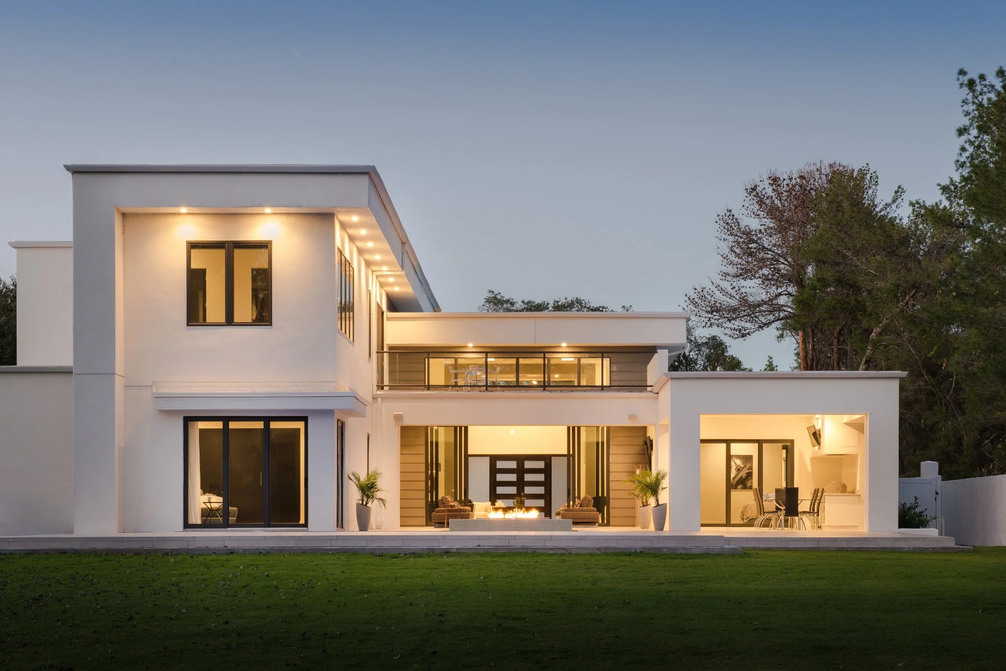 Modern white multi-story house with large glass windows and outdoor lighting, surrounded by a lawn and trees, during evening time.