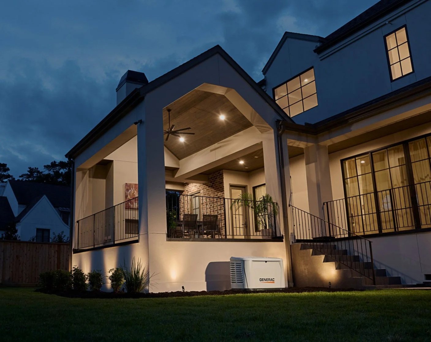 Modern two-story house at dusk powered by a Generac generator, with an illuminated outdoor patio, ceiling fan, and seating area.