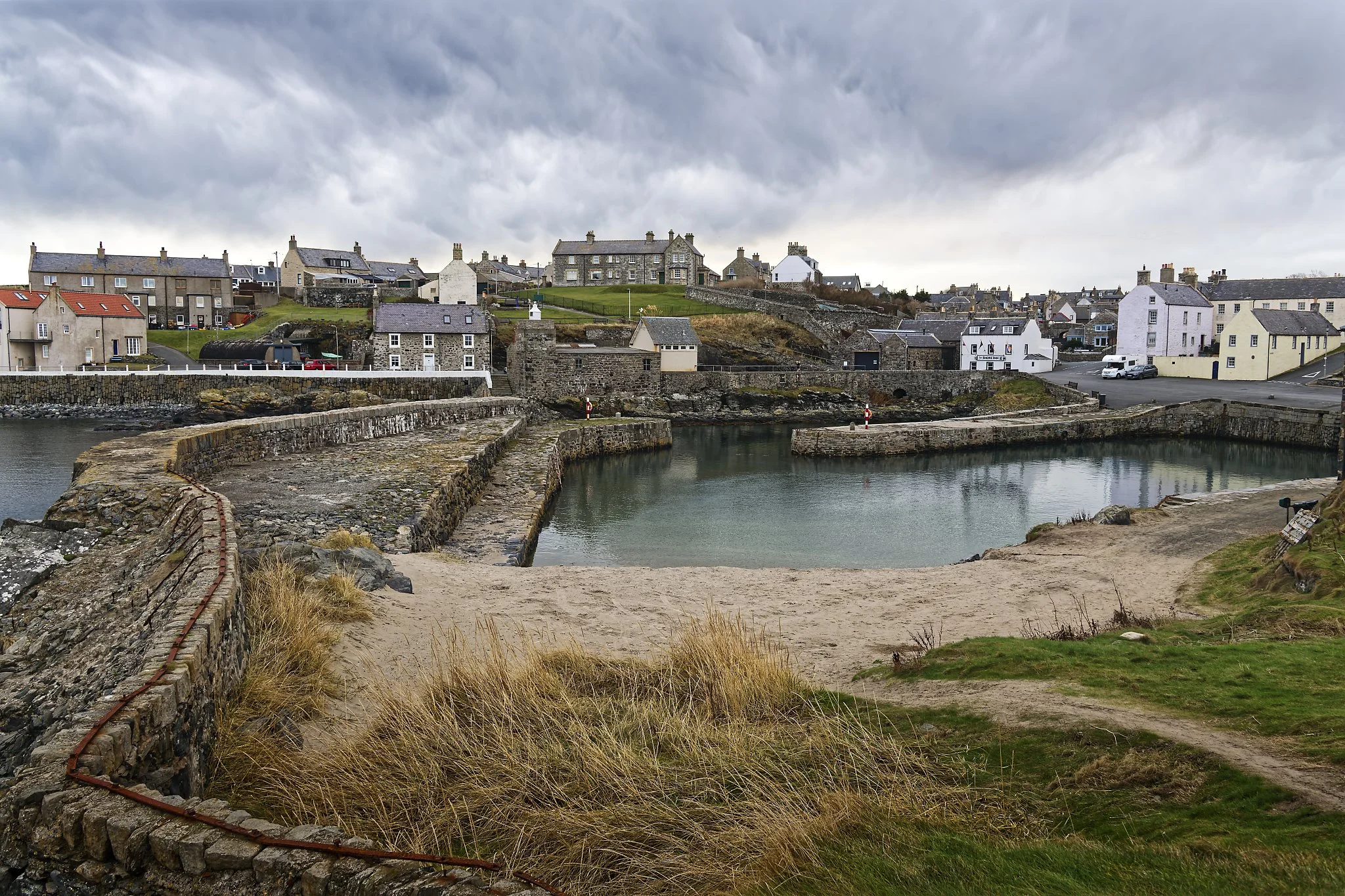 A coastal village with stone houses, a small harbor with stone breakwaters, and cloudy skies above. Portsoy Harbour  Scotland.
