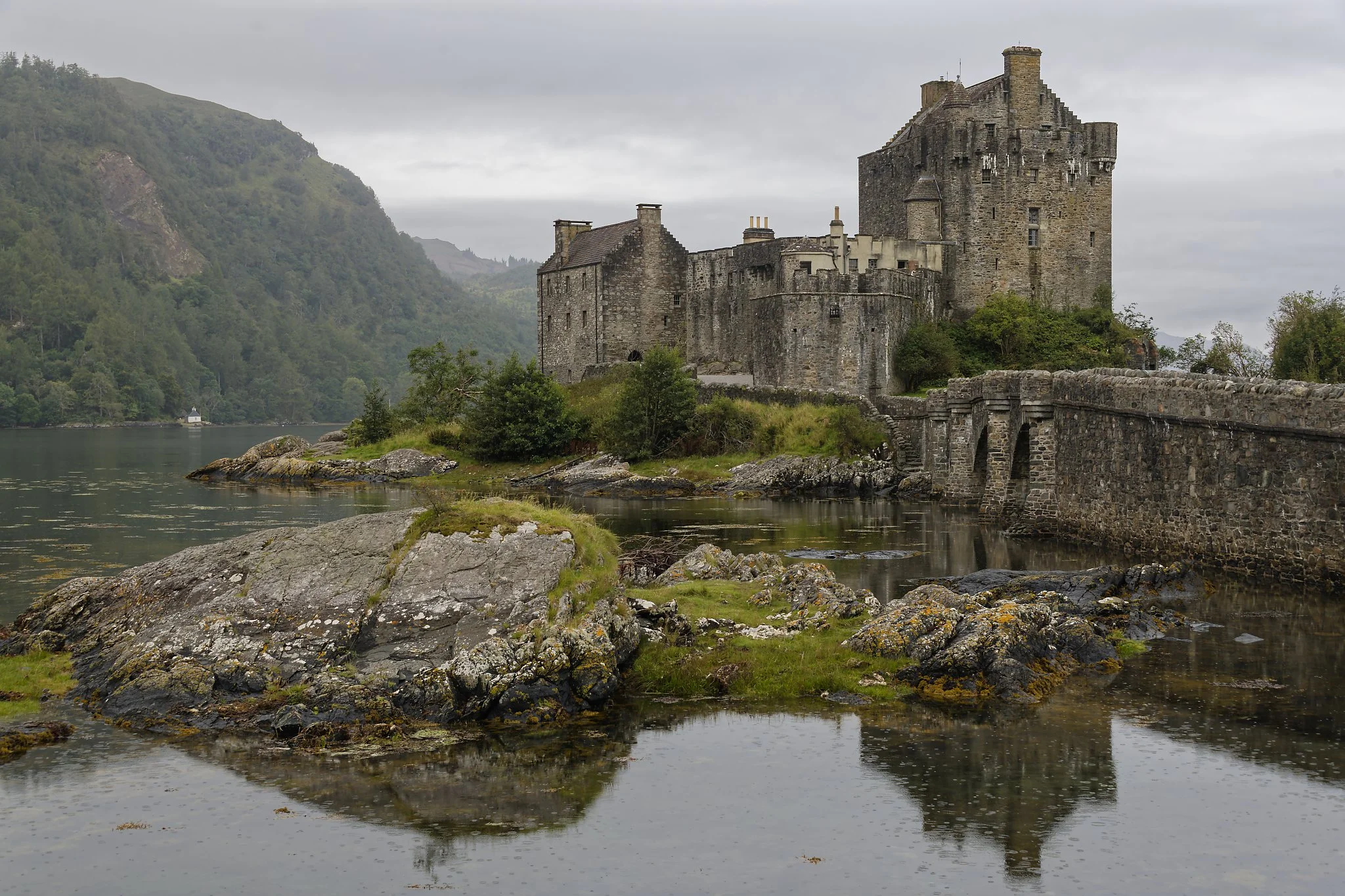 A historic stone castle situated on a small island in a lake, surrounded by lush green hills and overcast skies. Scotland