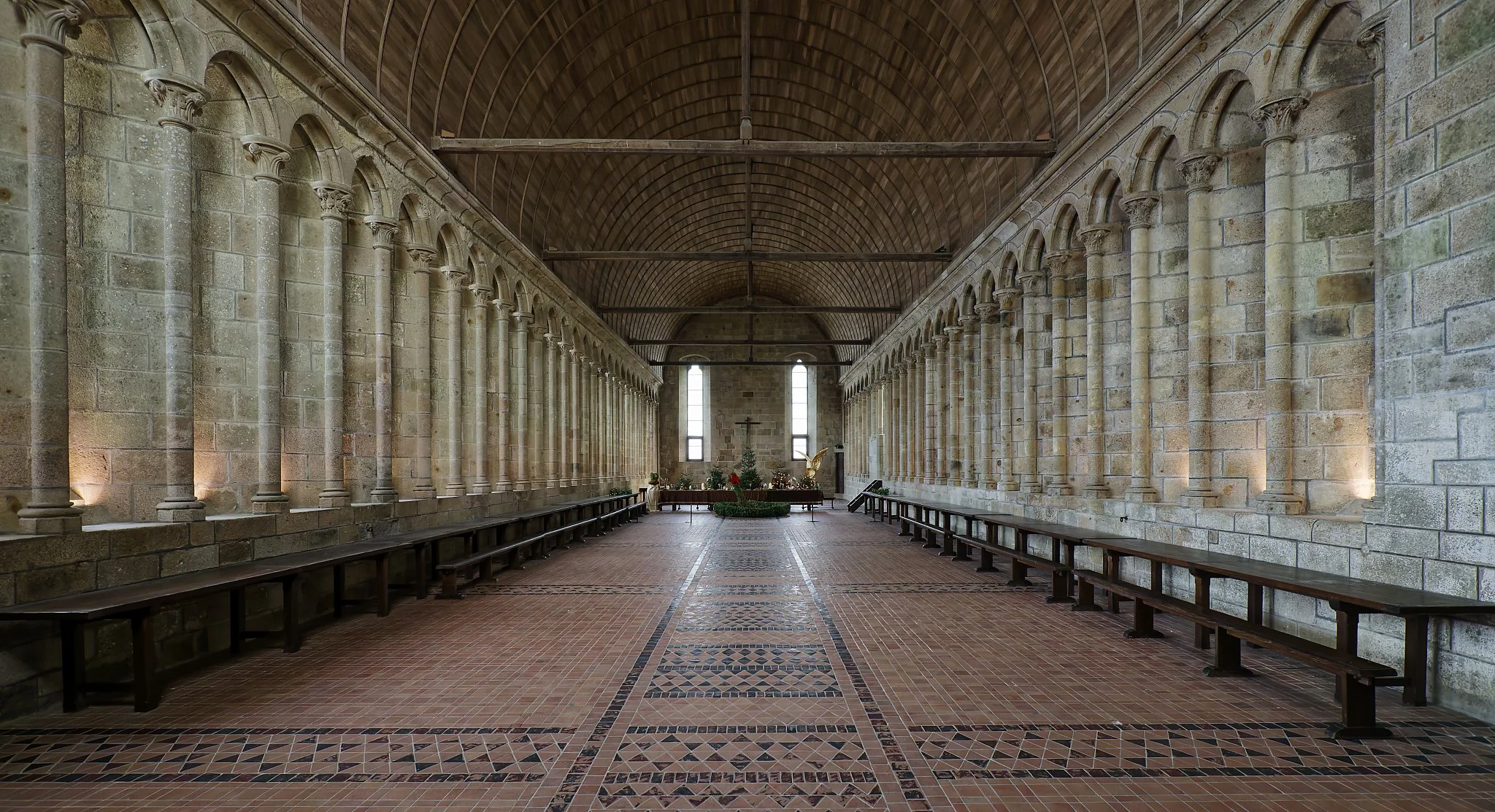 Interior of a church with stone walls, tall narrow windows, wooden ceiling, benches along sides, and an altar at the end with a cross and Christmas decorations.