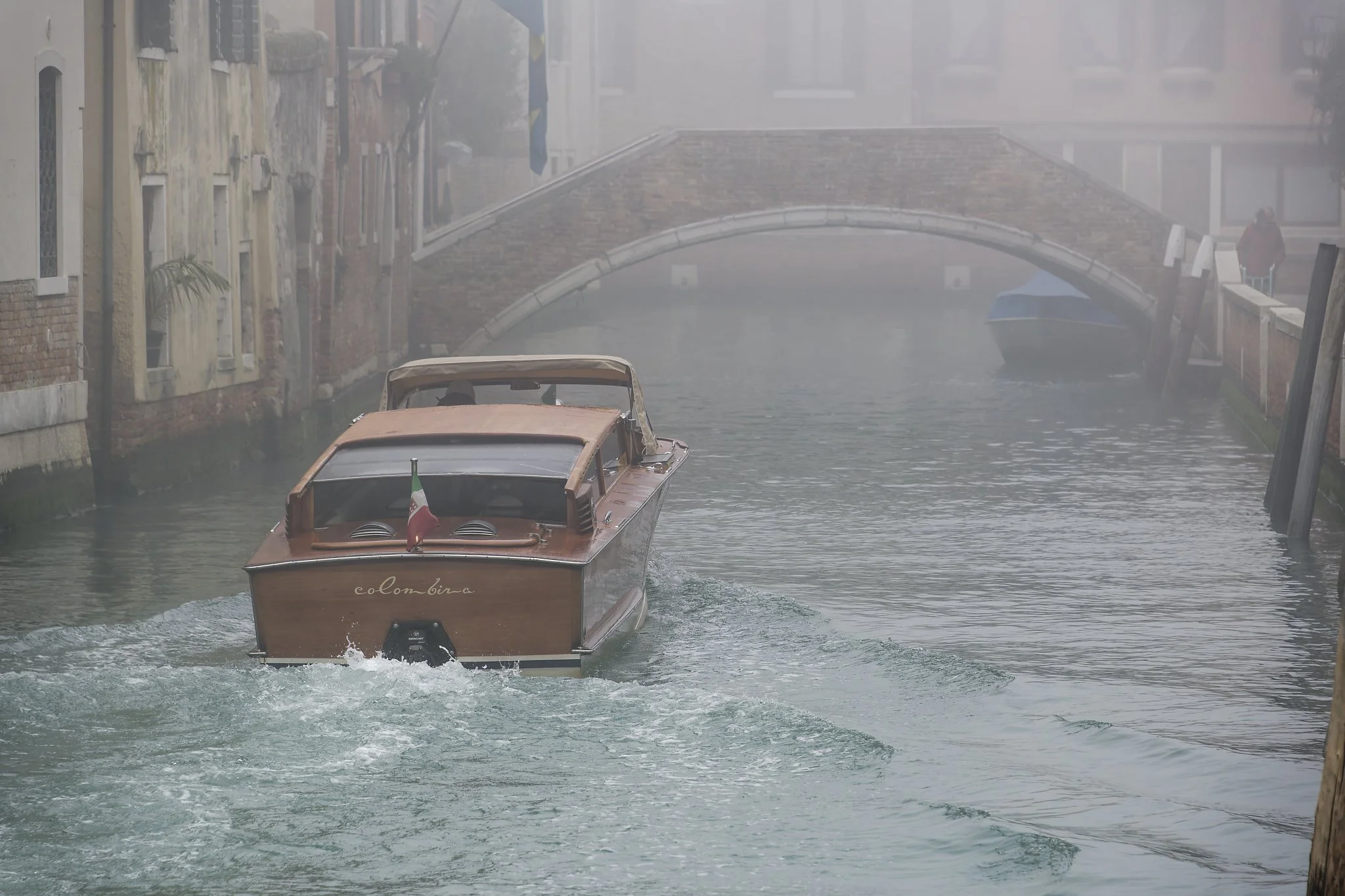 A wooden boat with an Italian flag is sailing through a canal with mist or fog, surrounded by buildings and a small stone bridge in Venice, Italy.