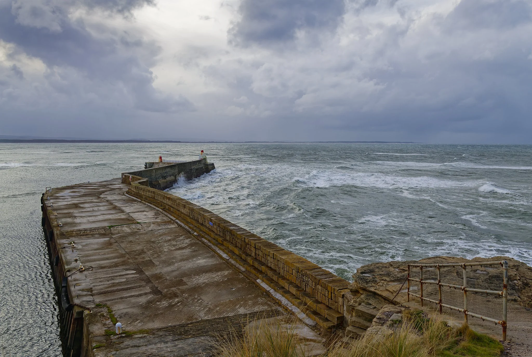 A concrete and brick pier extending into the ocean under a cloudy sky with storm clouds. Scotland.