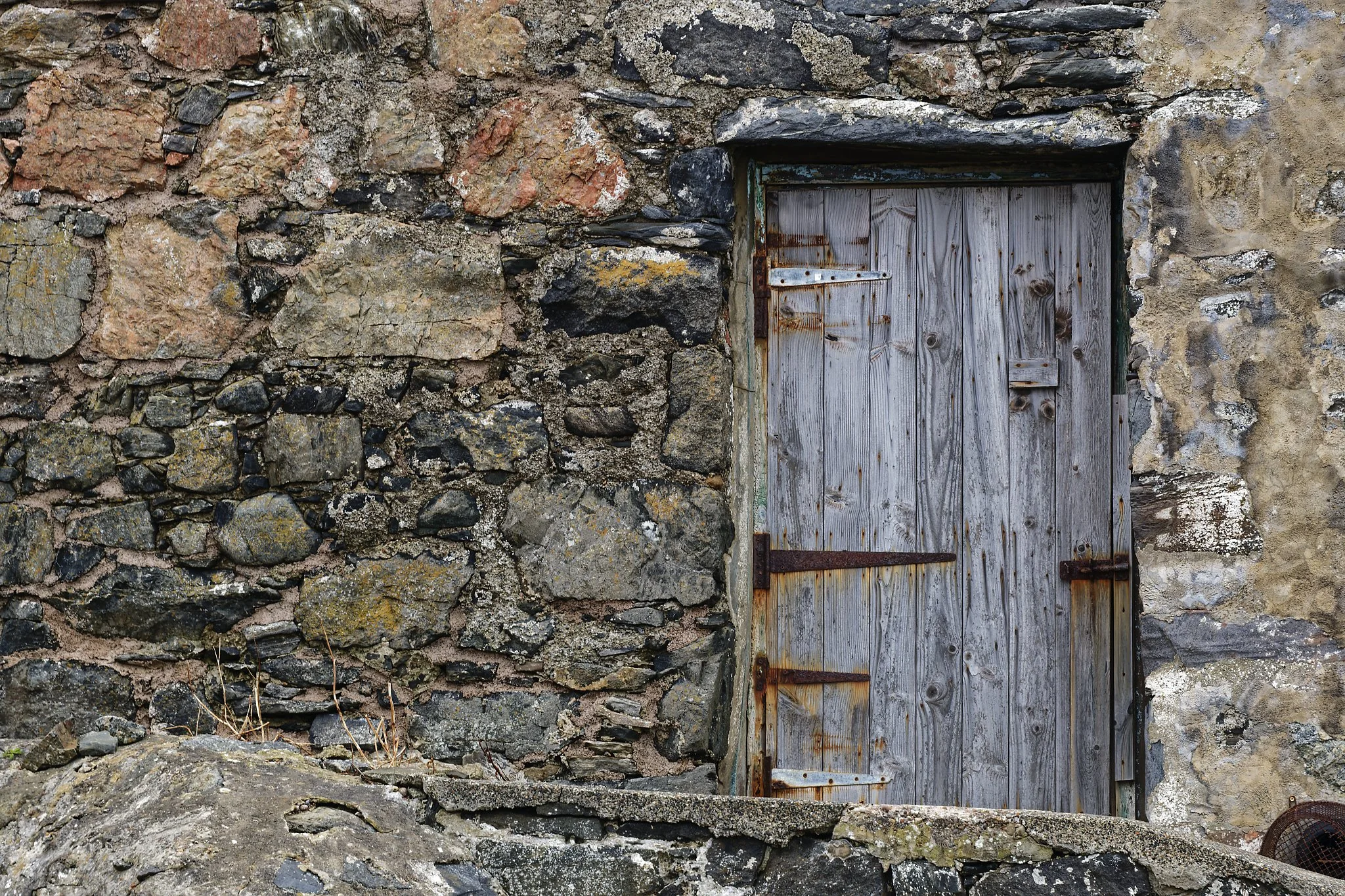 A weathered wooden door set into a stone wall. The door is grayish wood with rusted metal hinges and latch, surrounded by uneven stone and rock masonry.