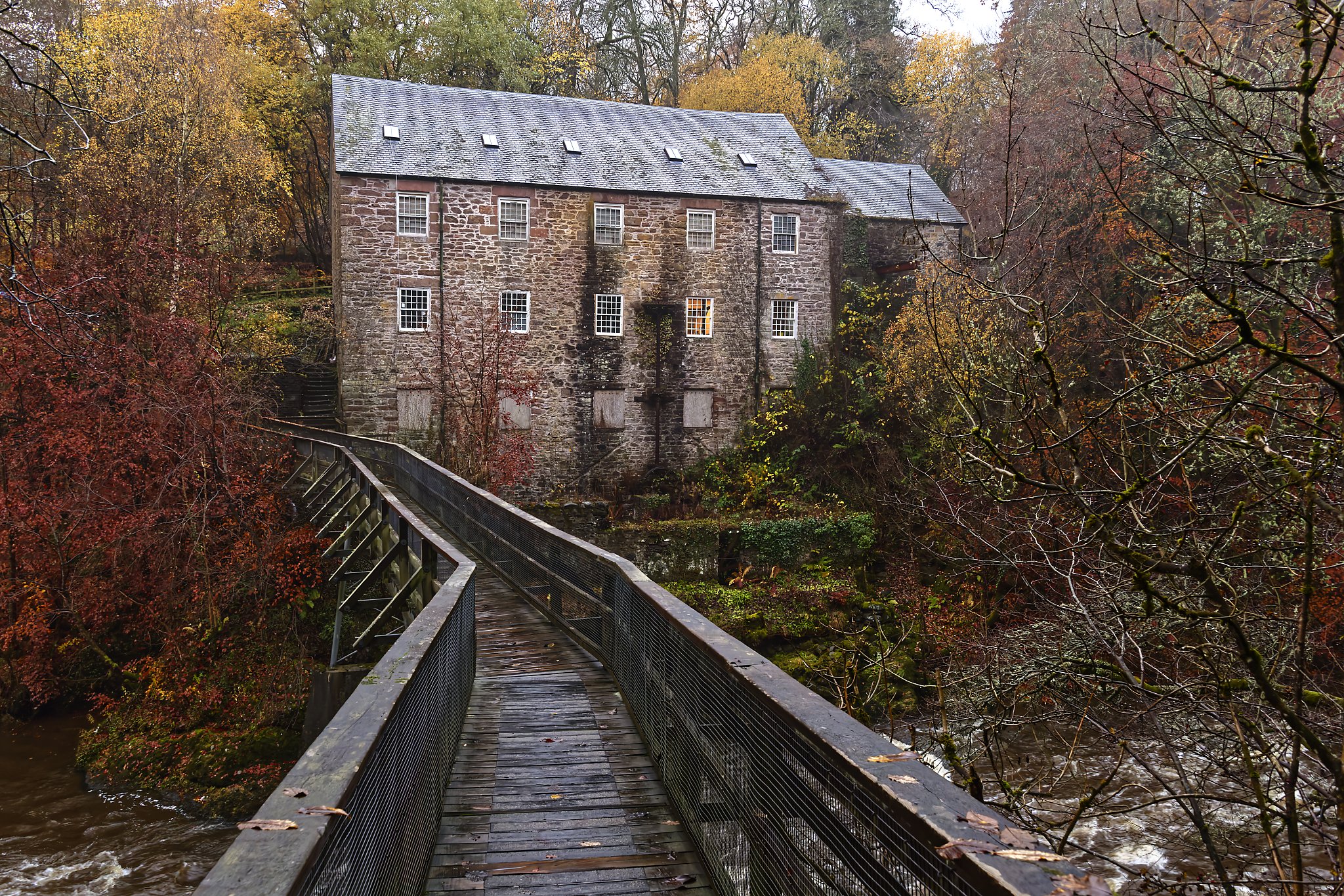 An old stone mill beside a river, connected by a wooden bridge, surrounded by autumn trees. Scotland.