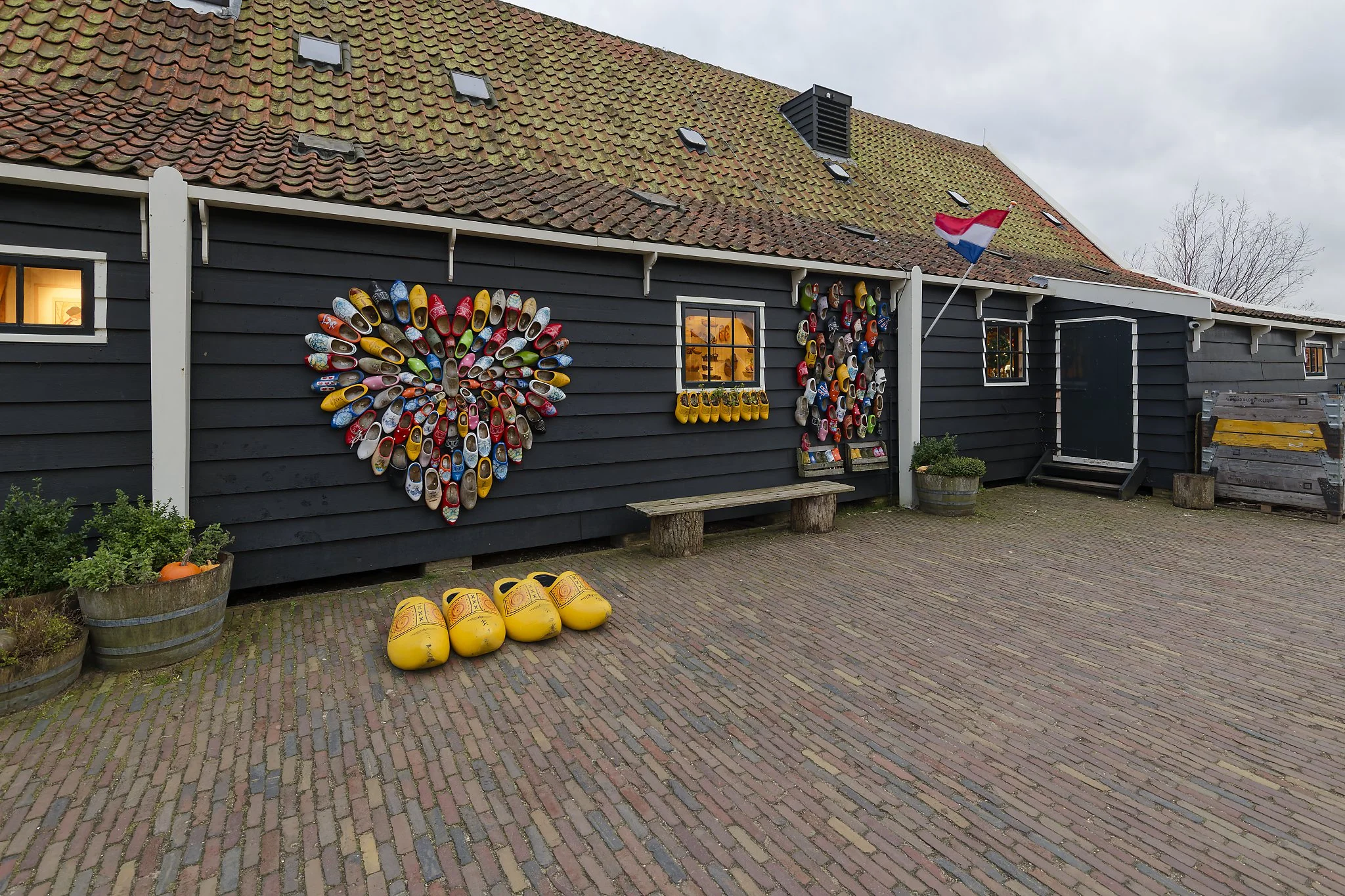 Wall decorated with colorful shoes arranged in heart and other shapes outside a rustic building, with wooden bench, potted plants, yellow oversized shoes, and a Dutch flag.