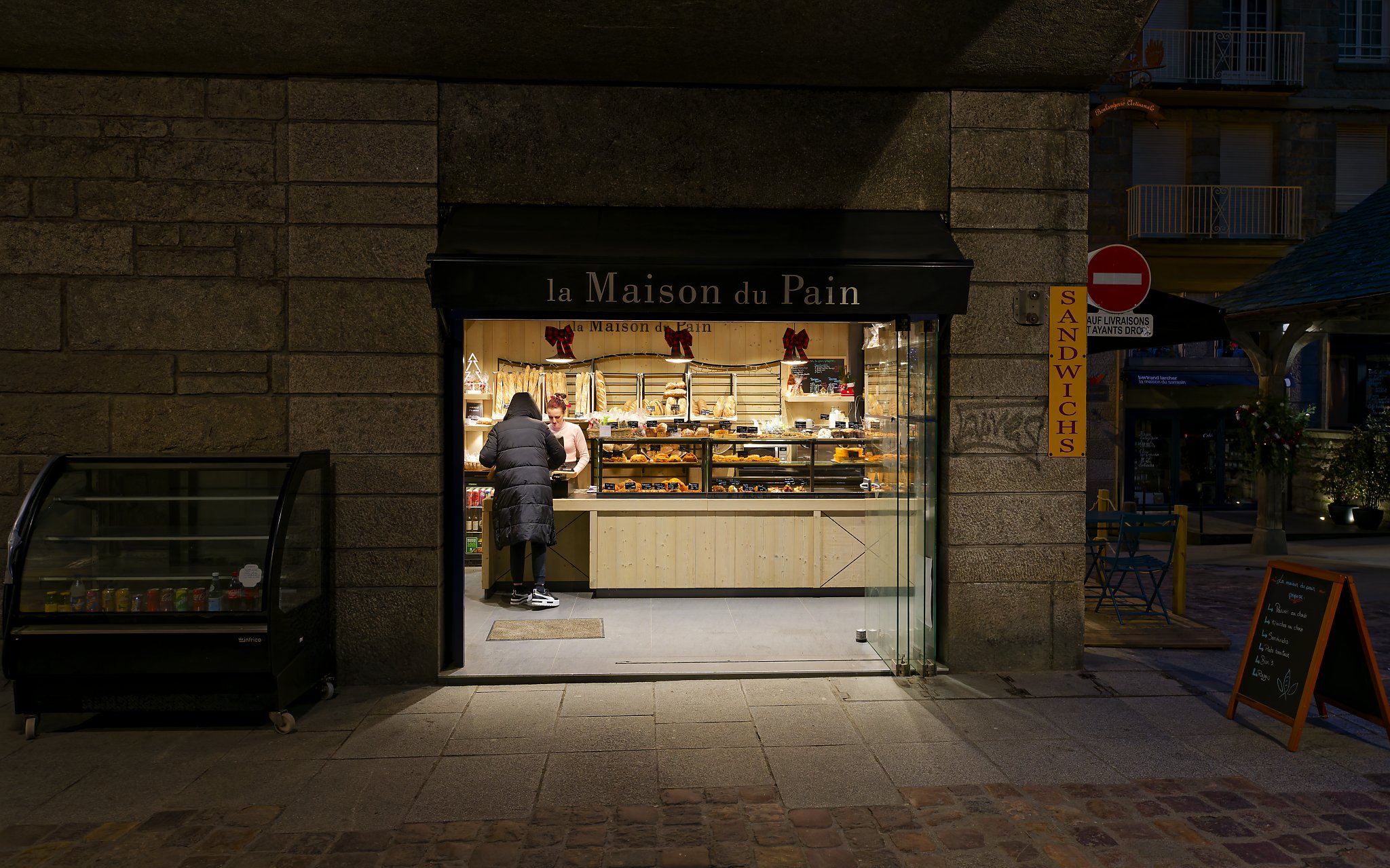 View of a bakery storefront with a sign that reads "la Maison du Pain" and decorated with red bows. Inside, a customer is ordering from a clerk behind the counter, with various baked goods displayed.