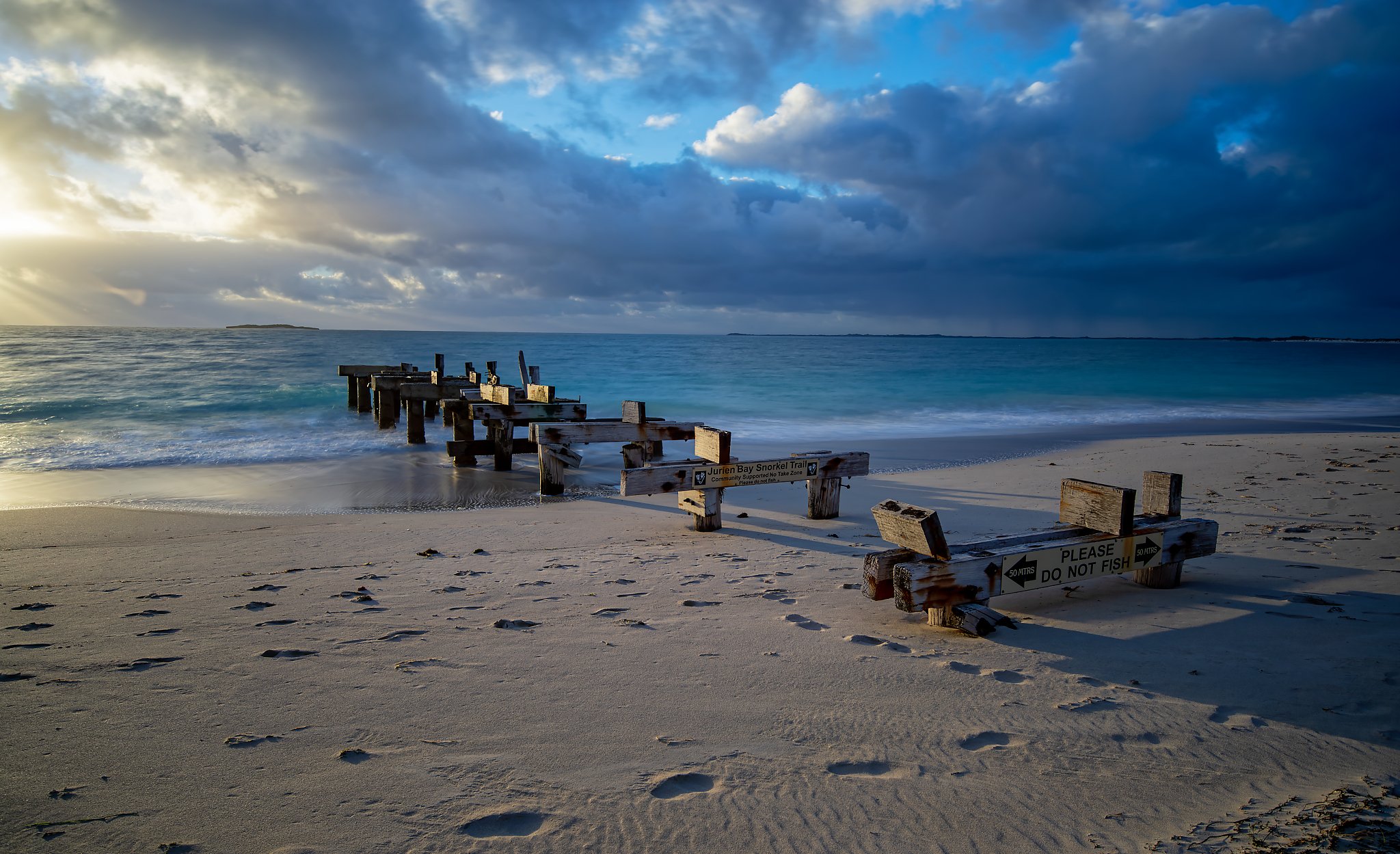 A beach with a wooden pier or barrier partly under water, with a sign that says 'Please Do Not Fish' and another sign indicating a 50-meter distance. The beach has footprints in the sand, and the ocean is calm with a cloudy sky overhead during sunset
