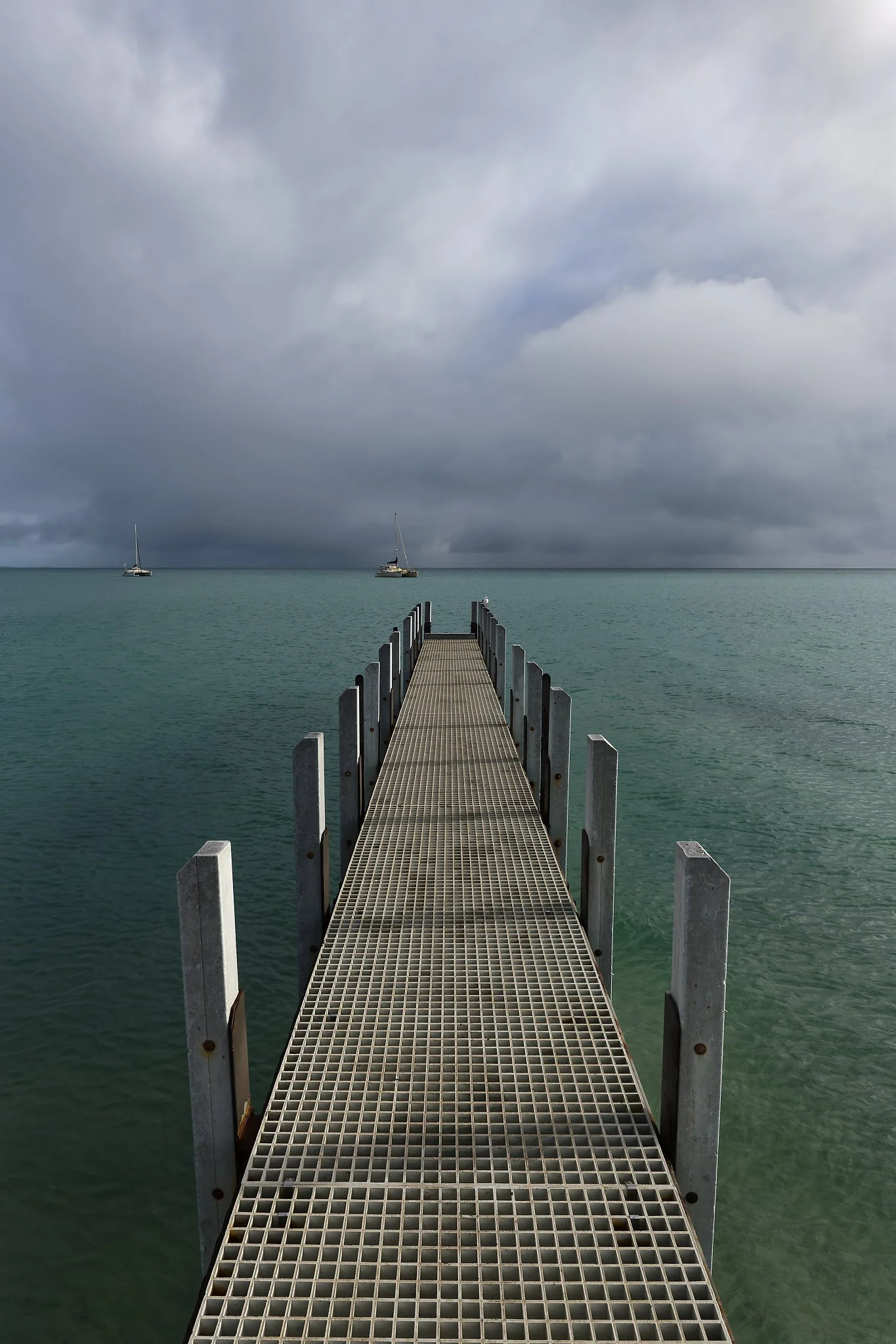 Metal dock extends into calm turquoise sea with two sailboats in the distance, under a cloudy gray sky. Exmouth Western Australia.