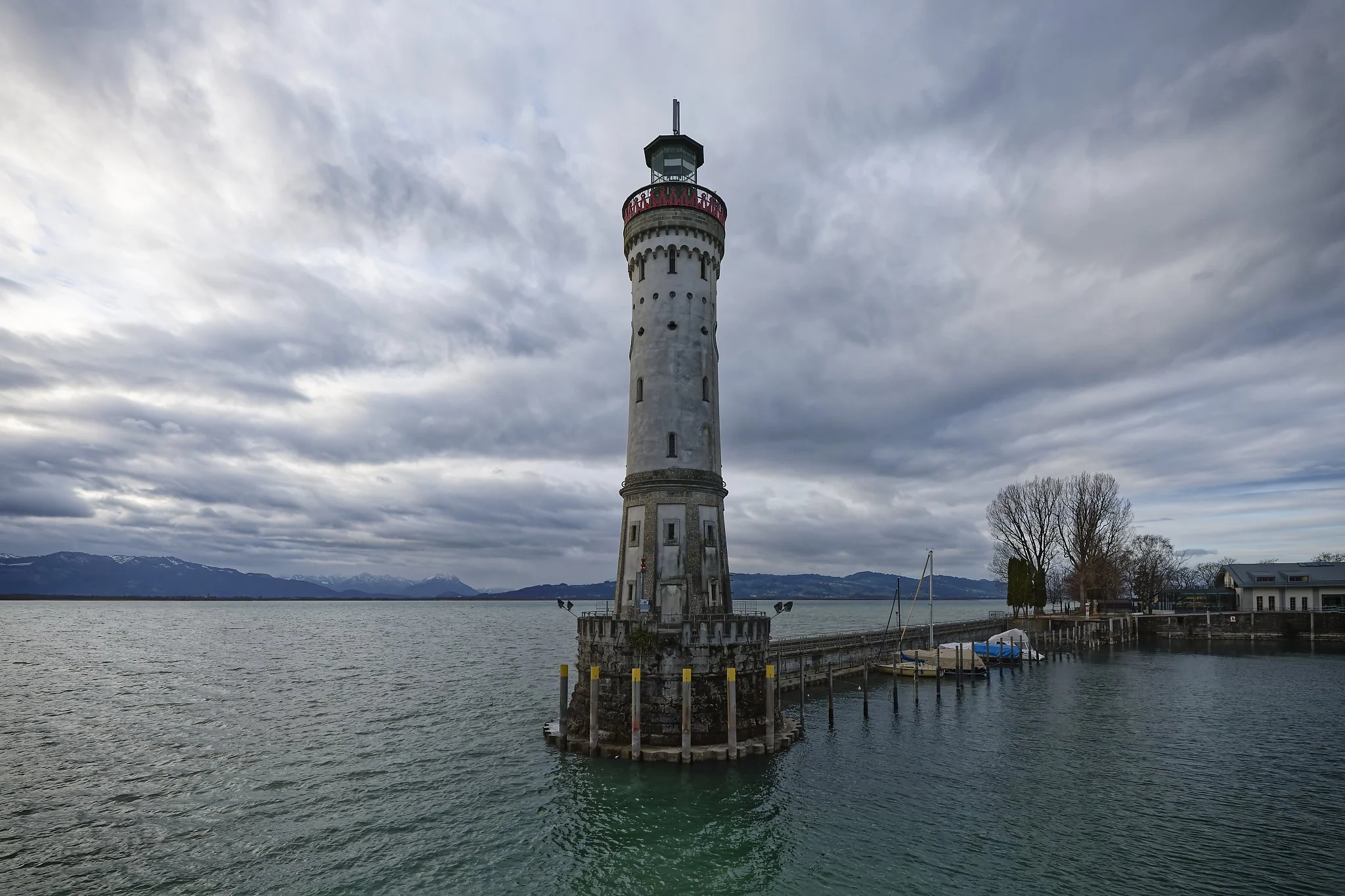 A lighthouse standing in the water near a dock with boats on the right, under a cloudy sky with mountains in the background. Germany.