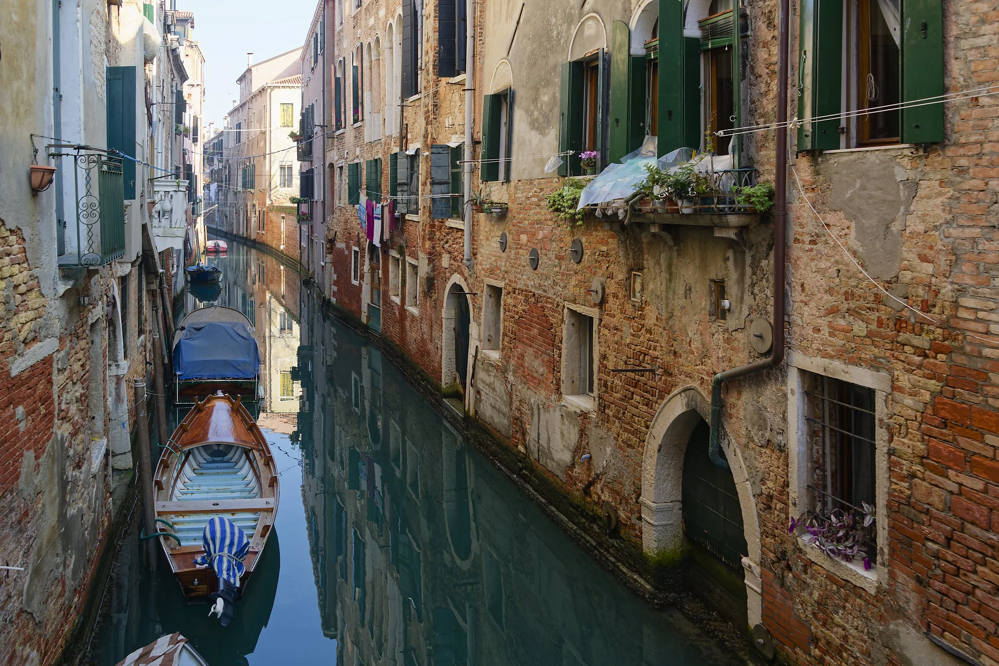 A narrow canal in Venice, Italy, with boats docked along the sides and old brick buildings with green shutters lining the waterway.