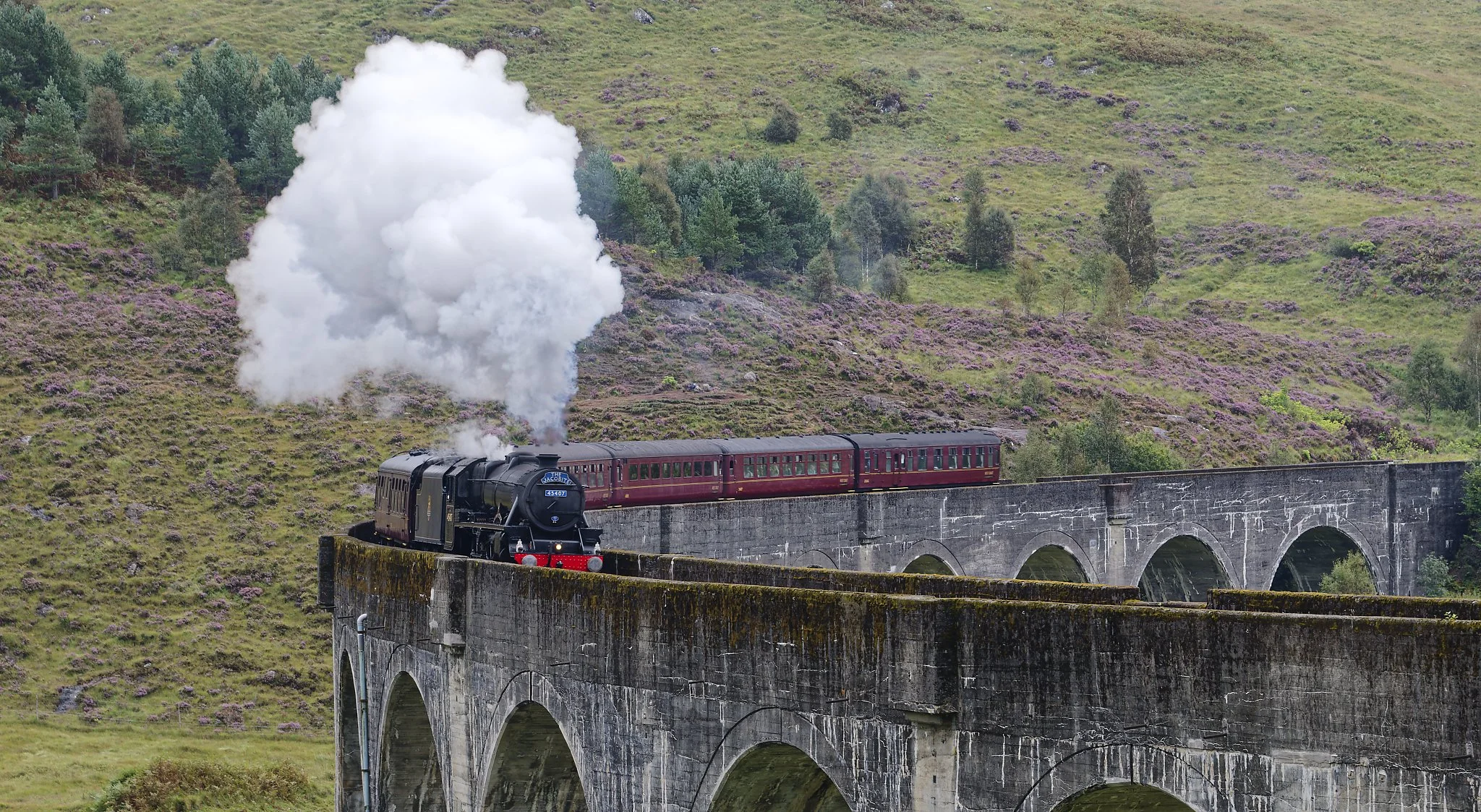 The Jacobite steam locomotive pulling red passenger cars crosses a stone arch bridge over a green hillside with purple heather and trees, with smoke billowing from the engine. Scotland.