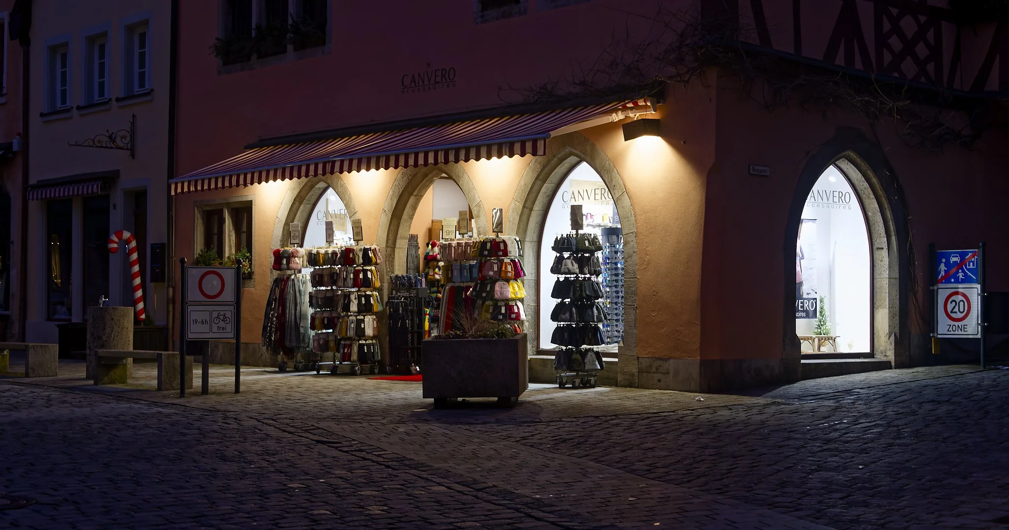 A boutique store named 'Canvero Accessories' illuminated at night, with display racks of handbags and accessories outside, situated in a European-style cobblestone street with signage indicating a bicycle zone and a speed limit of 20 km/h.