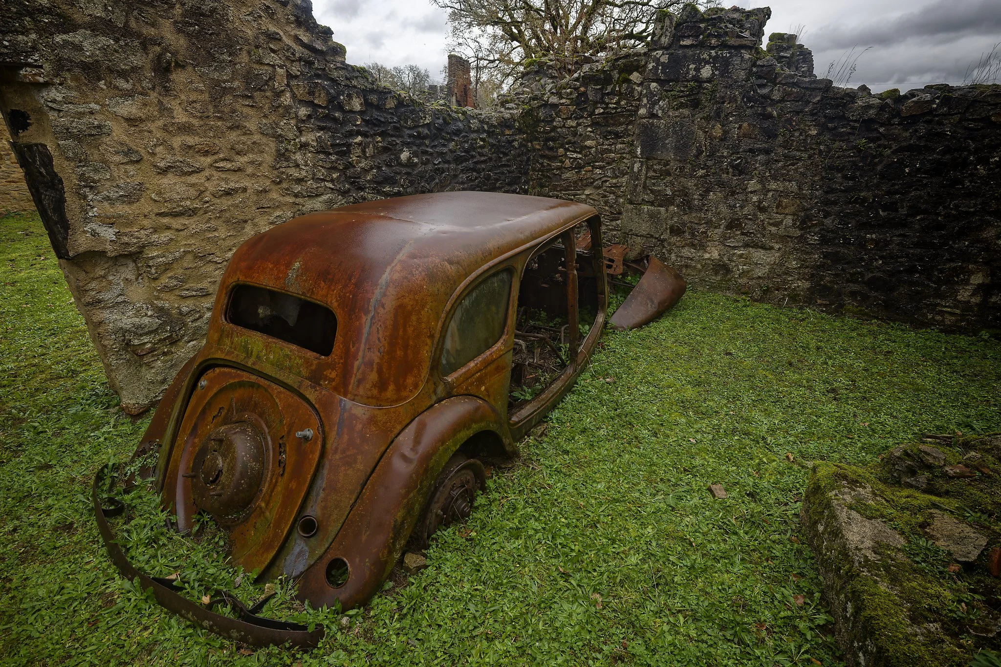 An old, rusted car without wheels, partially buried in green grass and surrounded by stone walls, under a cloudy sky. France.