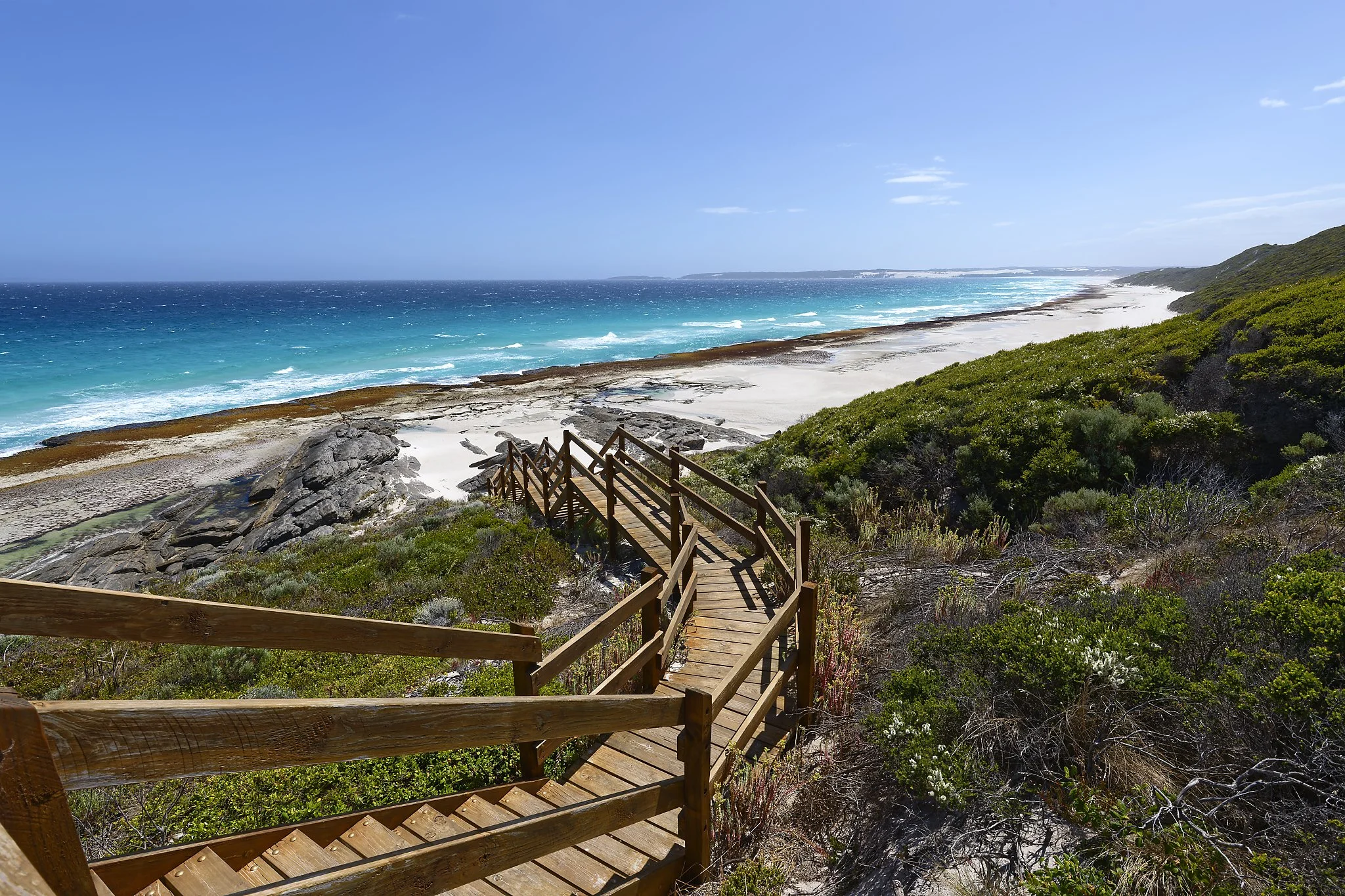 Wooden staircase leading down to a sandy beach with turquoise water and green shrubbery on hillsides under a partly cloudy sky. South Coast Western Australia.