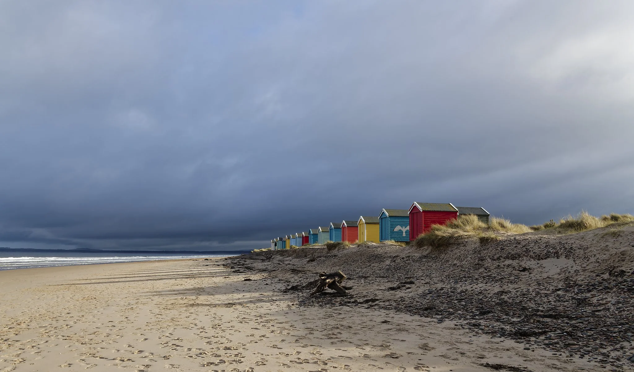 Colorful beach huts on a sandy beach with patches of grass, under a dark cloudy sky. Lossiemouth Scotland.