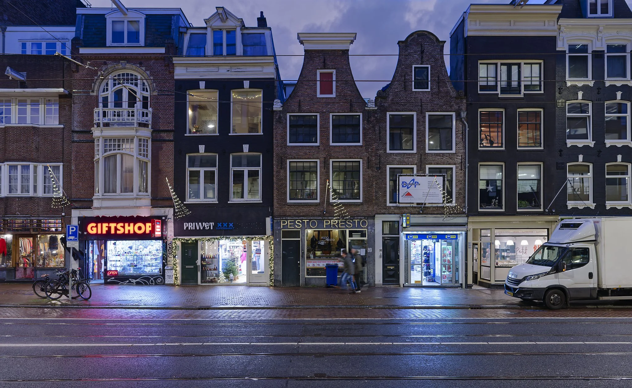 Nighttime city street with shops and apartments, lit with string lights, wet pavement, bicycles parked, and a white delivery van.