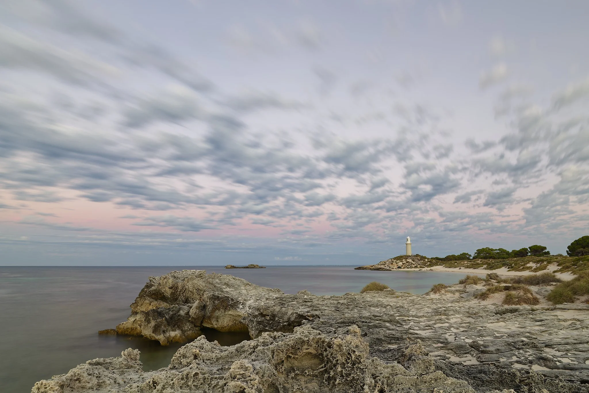 Scenic coastal landscape with rocky shoreline, a lighthouse on a distant hill, a calm sea, and a cloudy sky at sunset. Rottnest Island.