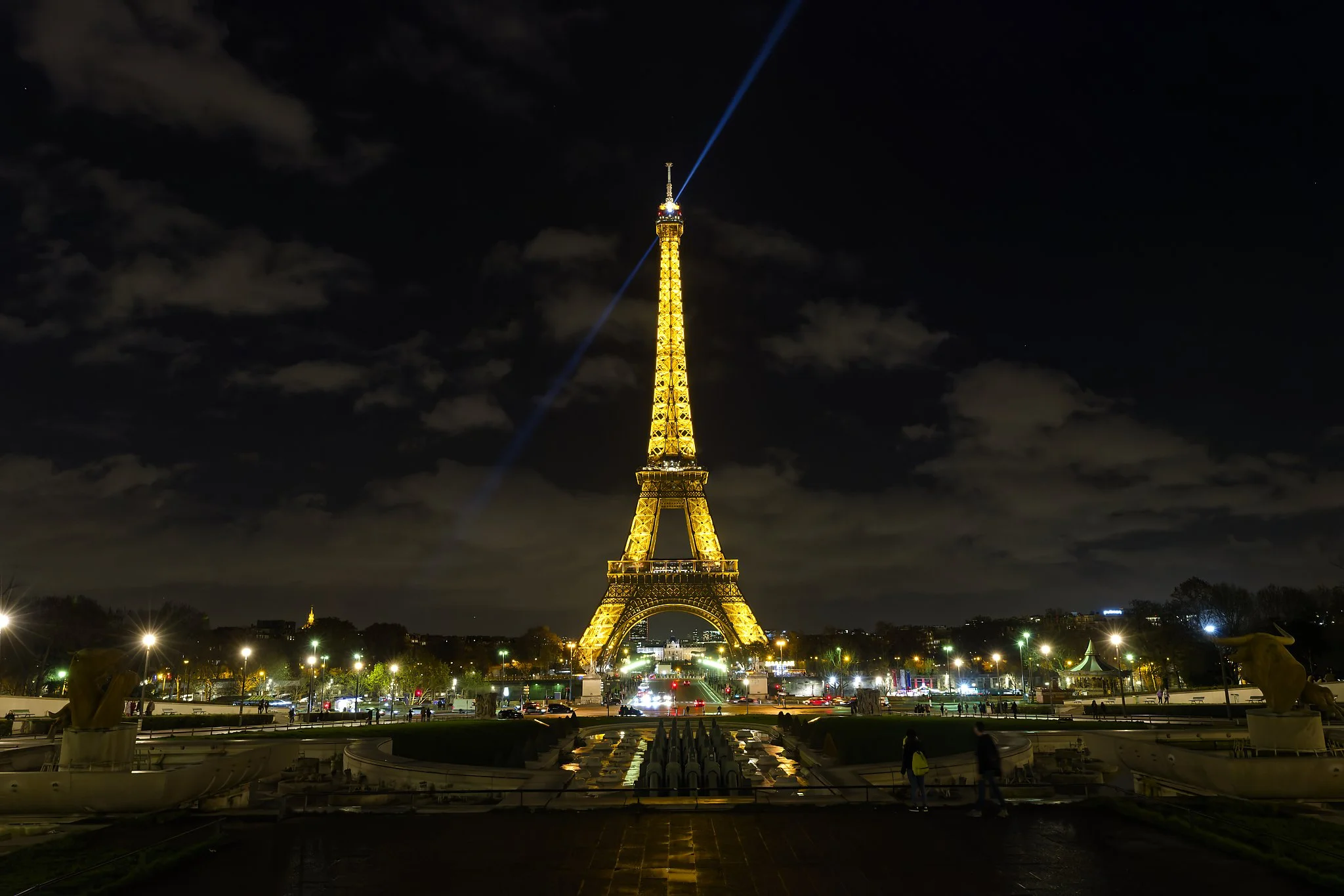 Night view of the Eiffel Tower illuminated in Paris with a spotlight beam pointing upwards, street lights and fountains in foreground, dark cloudy sky in background.