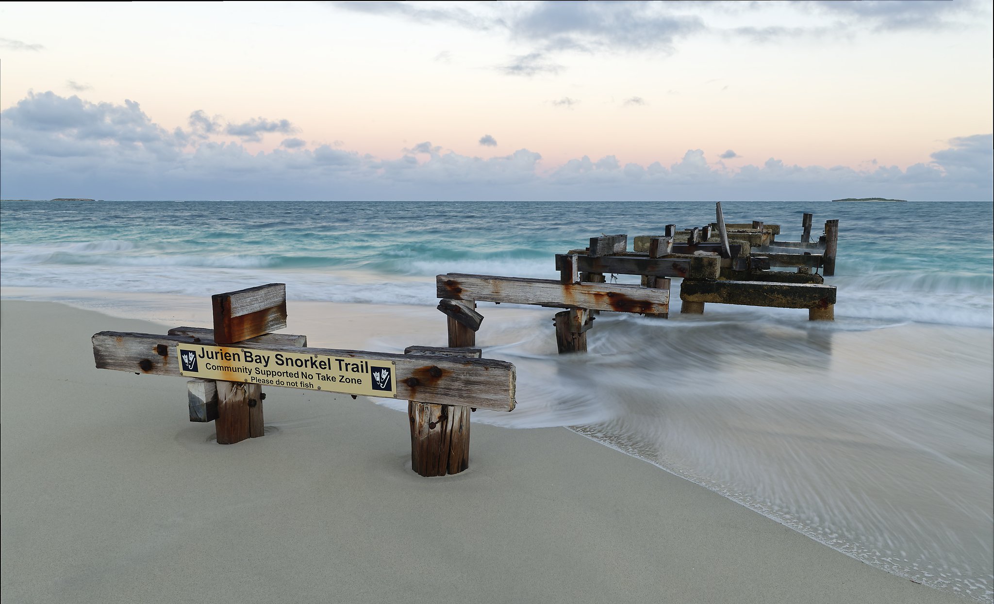 Wrecked wooden pier on a sandy beach with water and waves, sign reading 'Jurien Bay Snorkel Trail, Community Supported No Take Zone, Please do not fish'.