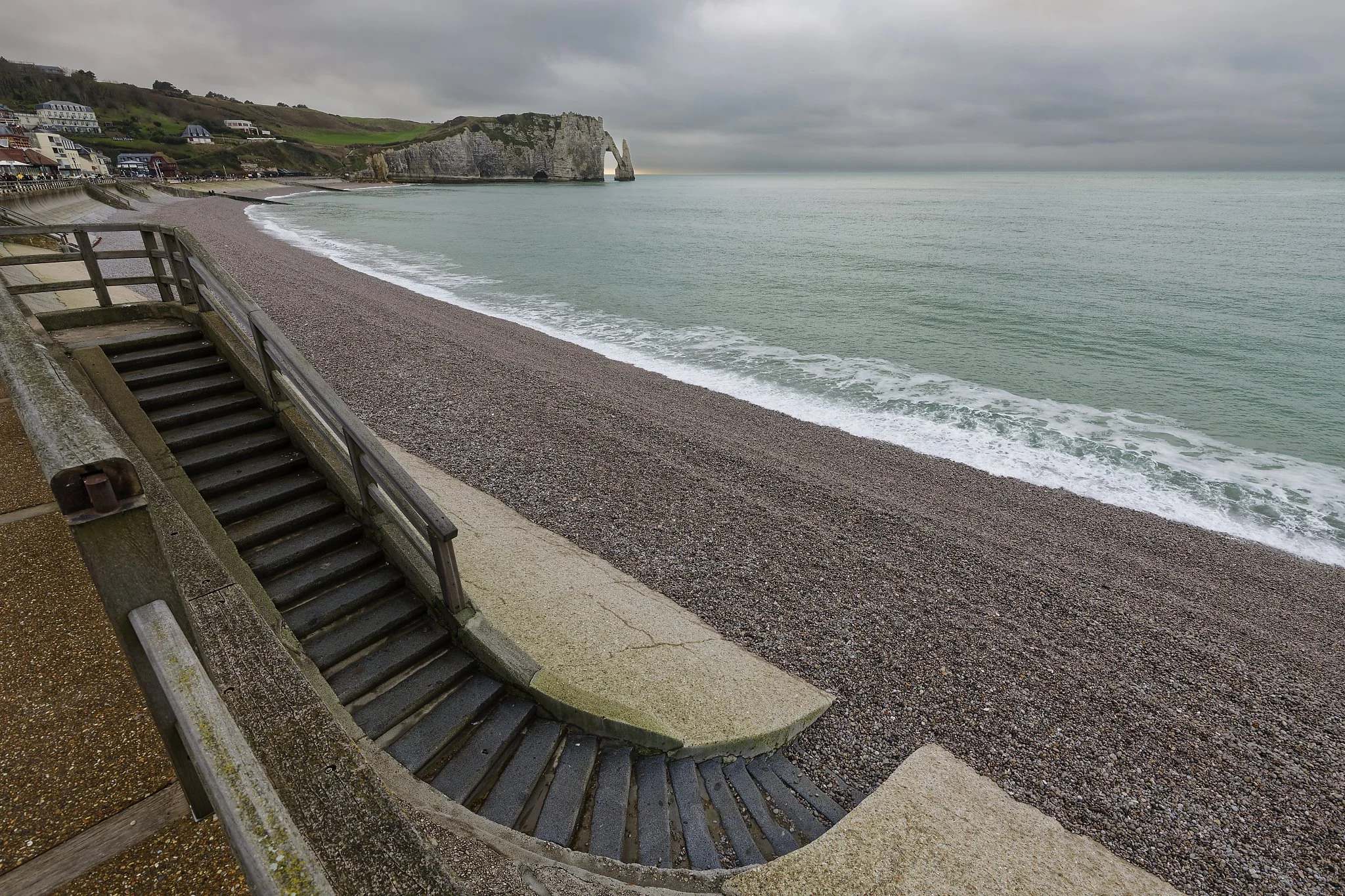 View of a pebble beach with a staircase leading down to the shore, overlooking the ocean, with cliffs and houses in the background on a cloudy day.
