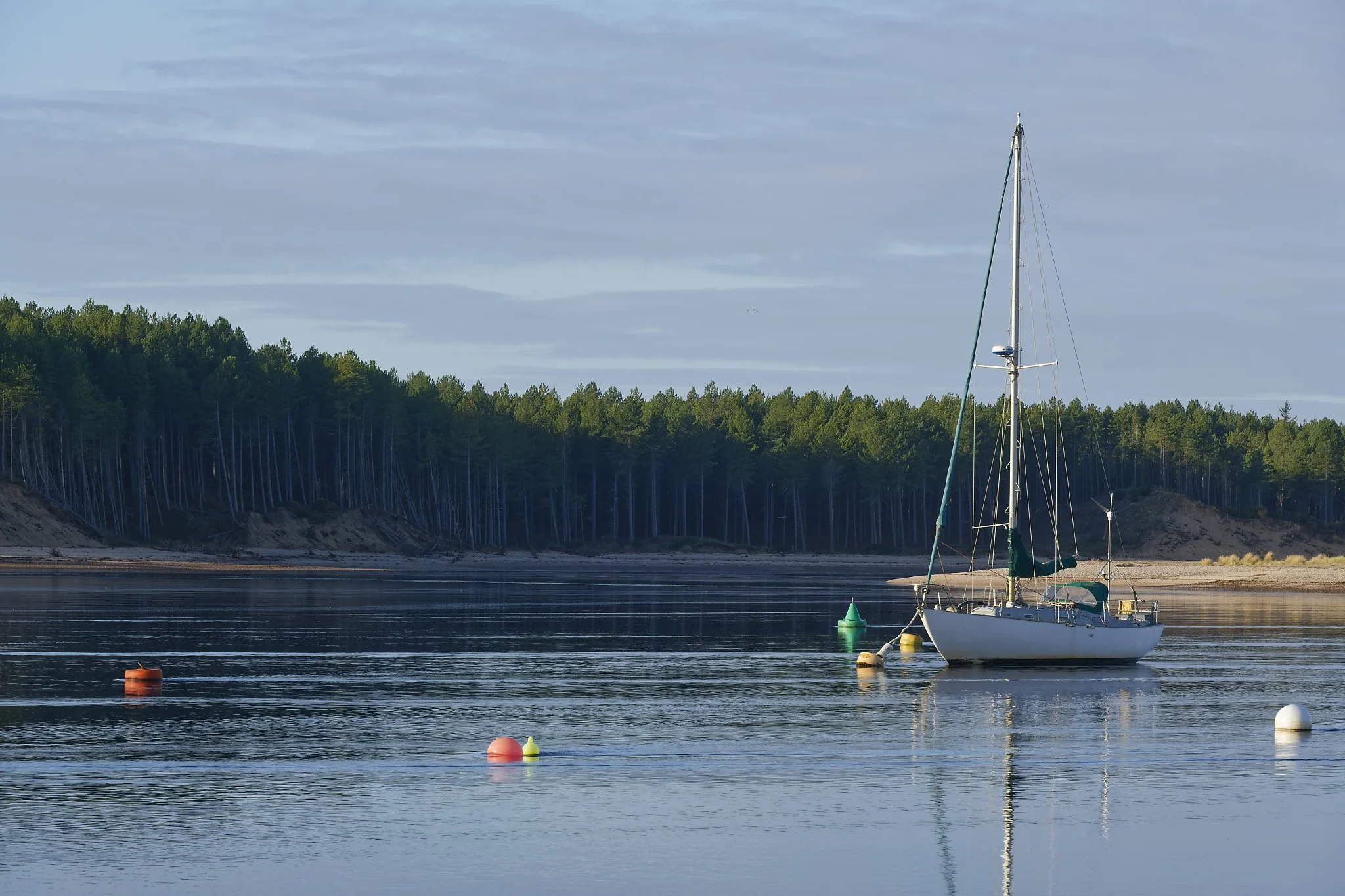 A sailboat anchored in calm water near buoys with a tree-lined shoreline in the background. Lossiemouth Scotland.