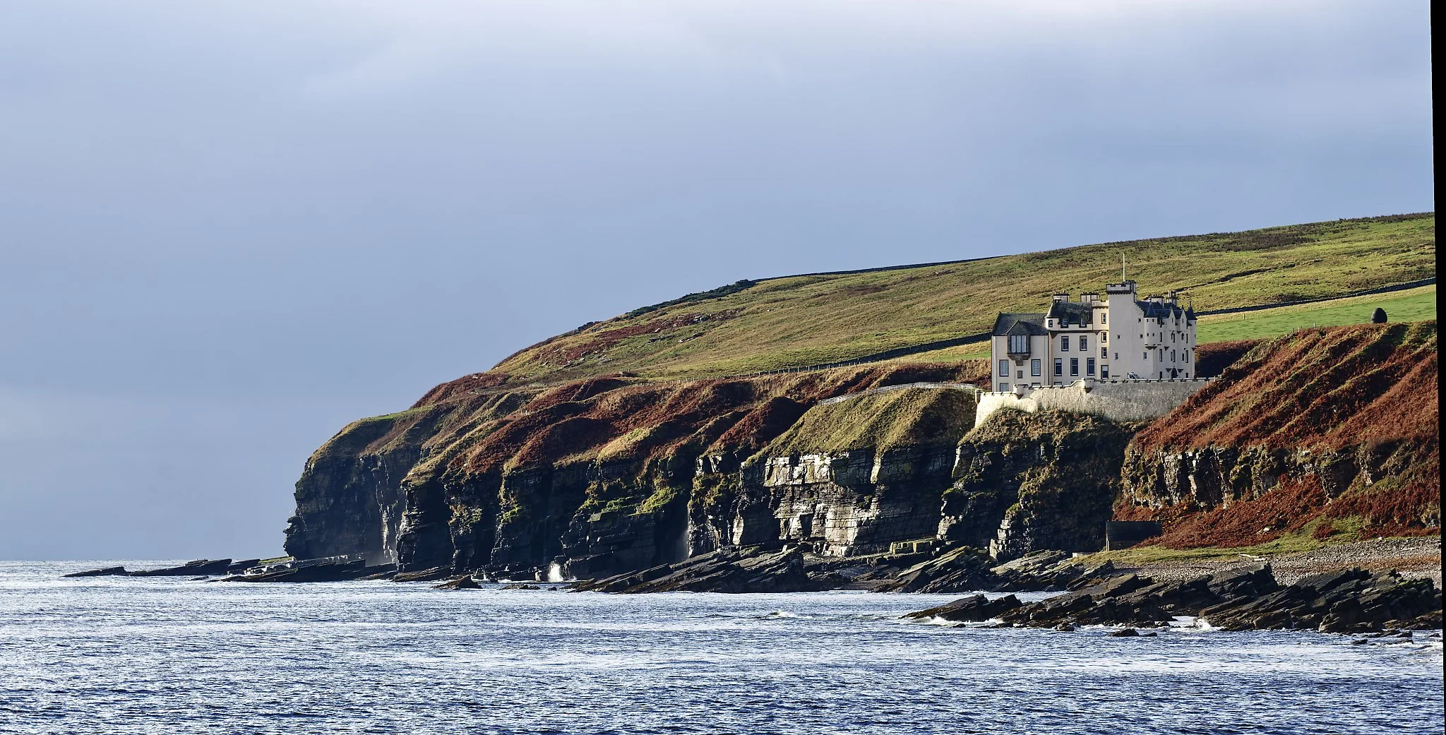 A large white castle-style house located on a grassy cliff overlooking the ocean, with rocky cliffs below and a cloudy sky above. Scotland.