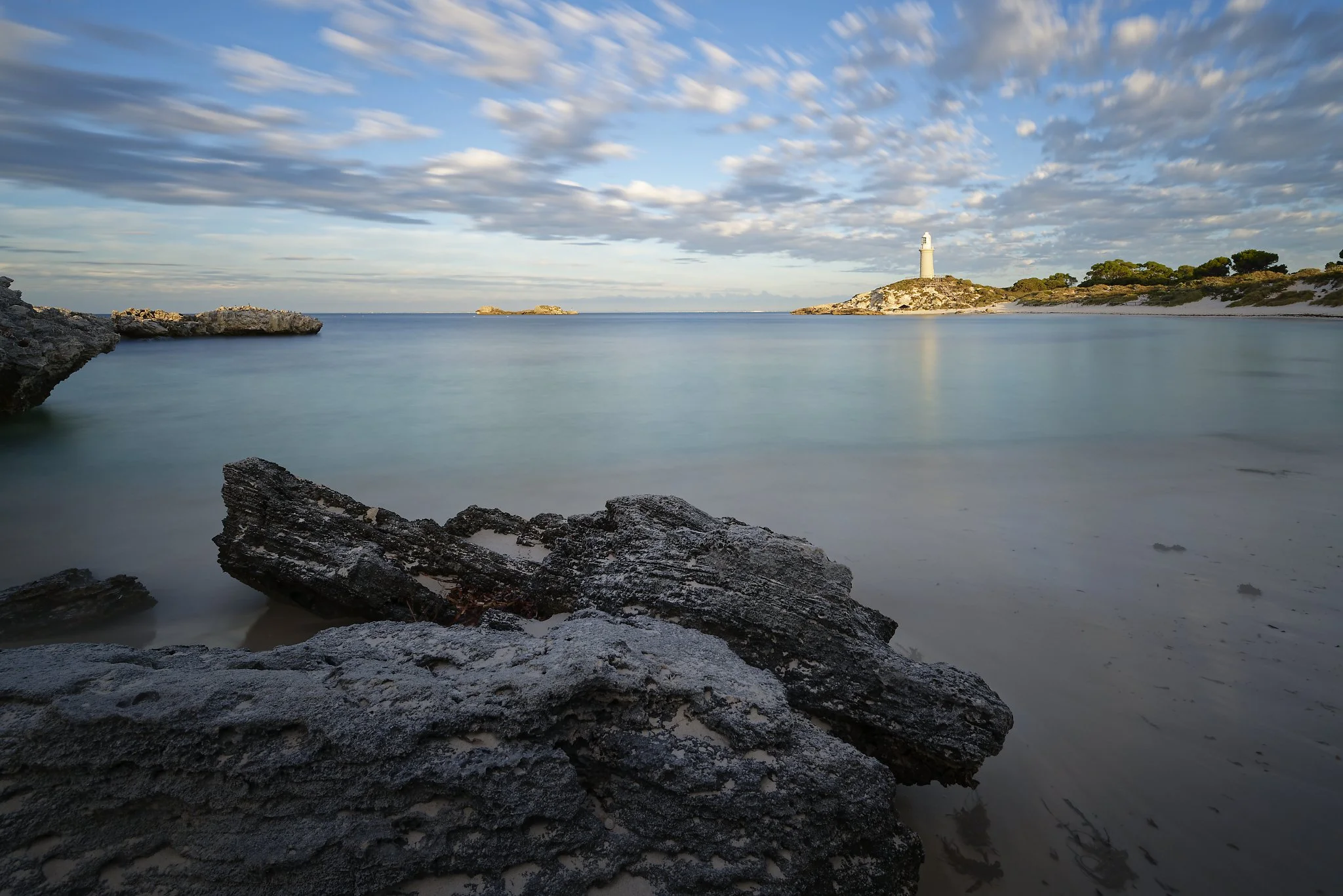Long exposure shot of a calm ocean bay with rocky shoreline in the foreground, a lighthouse on a hill in the distance, and partly cloudy sky. Rottnest Island Western Australia.