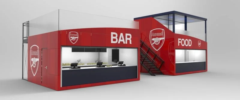 A modern, two-story food stall with red exterior walls featuring Arsenal Football Club logos. The lower level has a bar counter, and the upper level has a food service window accessed by exterior stairs.