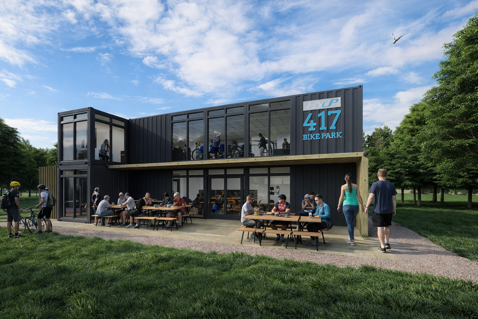 People gathering outdoors at the 417 Bike Park in a building made of black shipping containers with large glass windows, with some riding bikes inside and others sitting and walking outside.
