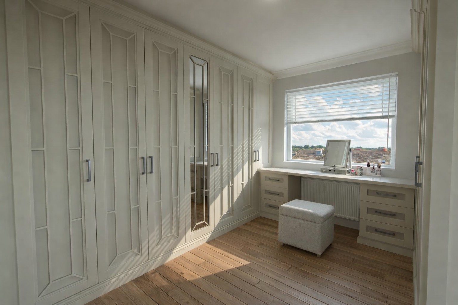 A bright room with white built-in wardrobes, a white desk with a mirror, a small cushioned stool, and a large window with blinds showing a view of the sky and rooftops.