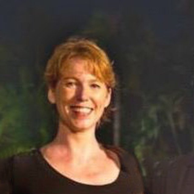 A smiling woman with short red hair wearing a black top standing outdoors at night with trees in the background.