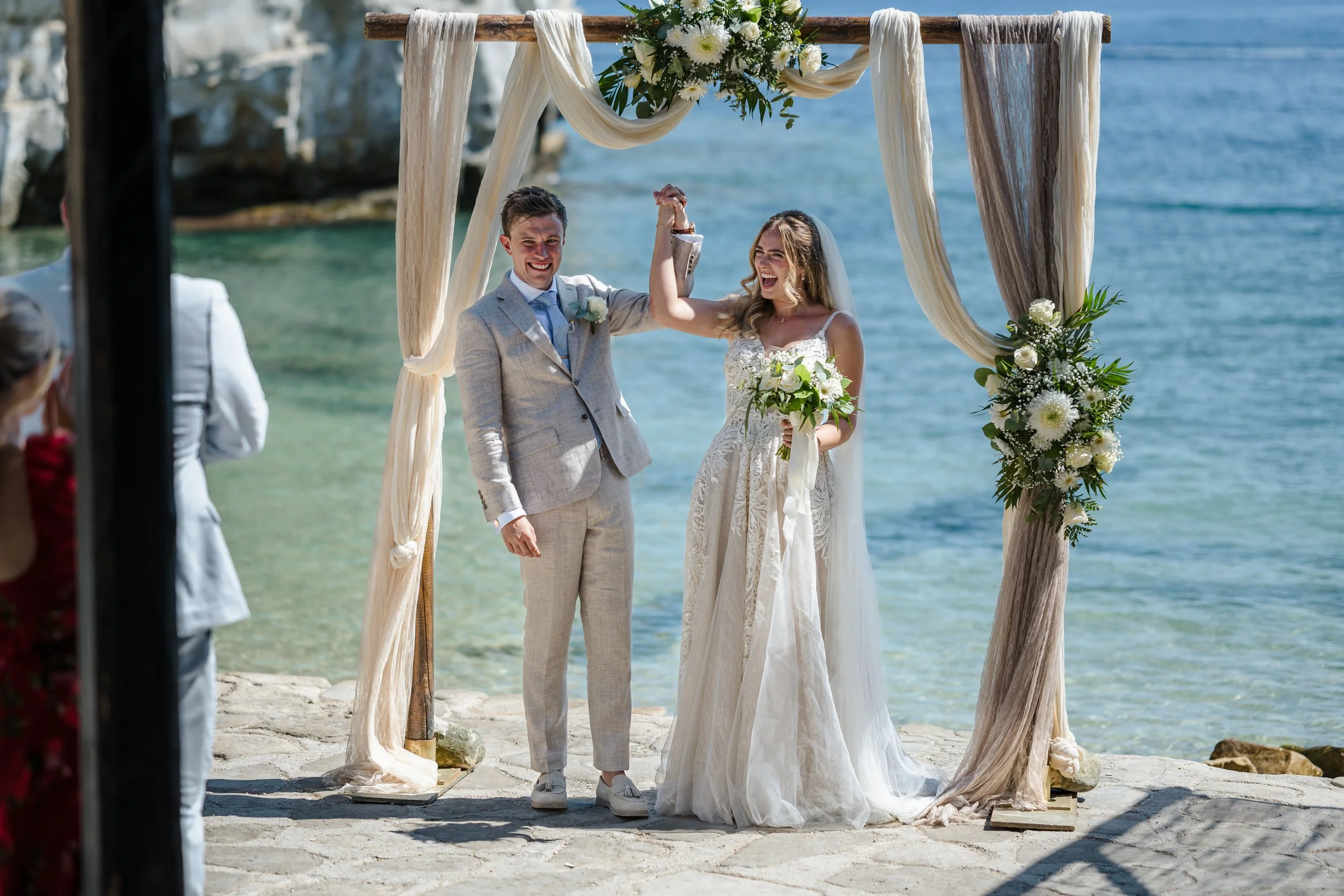 A newly married couple standing under a wooden wedding arch decorated with white flowers and drapery on a beach, celebrating their wedding with the ocean in the background. The bride is holding a bouquet and smiling, while the groom is smiling and holding the bride's arm. Guests are visible in the foreground.