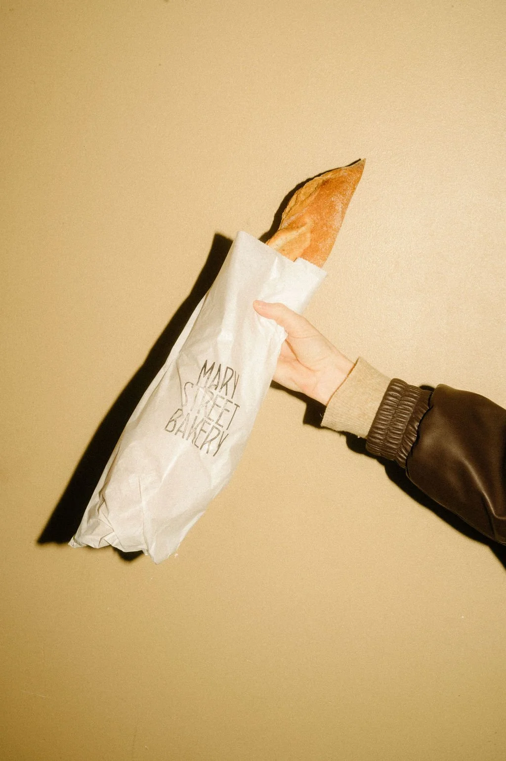 A person holding a baguette partially wrapped in a paper bag from Mary Street Bakery against a beige background.