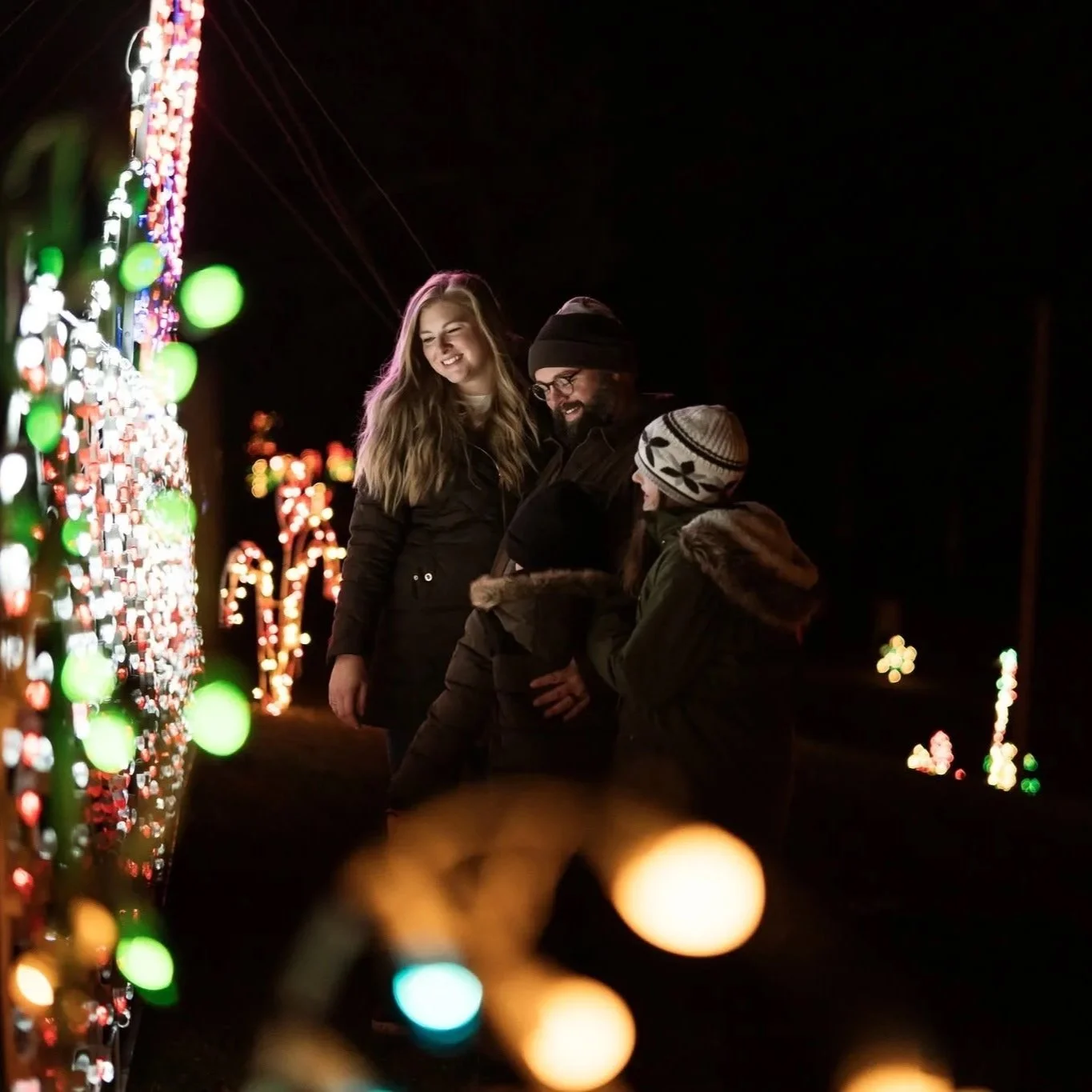 A family of four enjoying Christmas lights at night, standing together and smiling. They are dressed warmly for winter, with colorful holiday lights in the background.