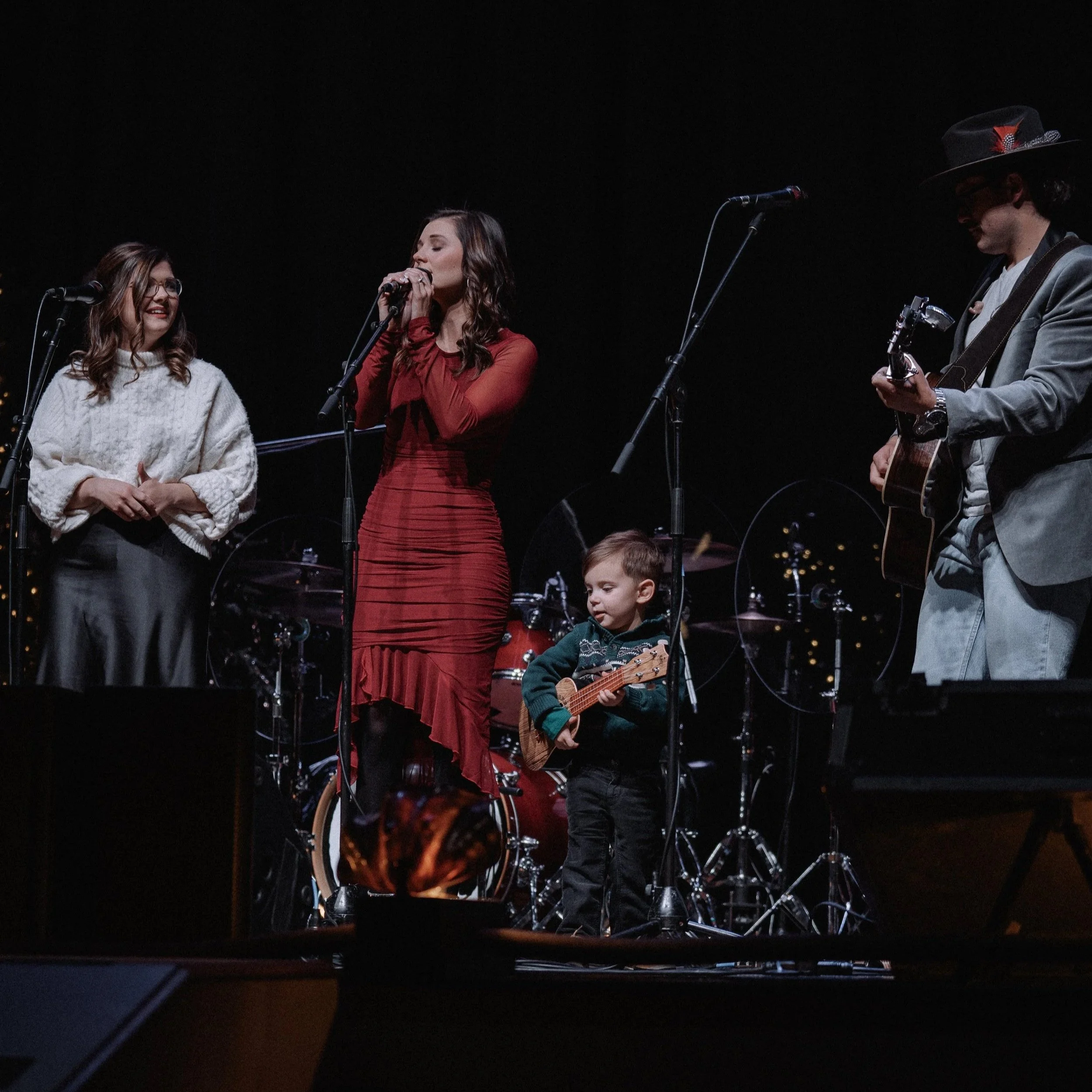 Three people performing music on stage at night, including a woman singing with closed eyes, a woman smiling at her, and a man playing guitar wearing a hat, with drums in the background.